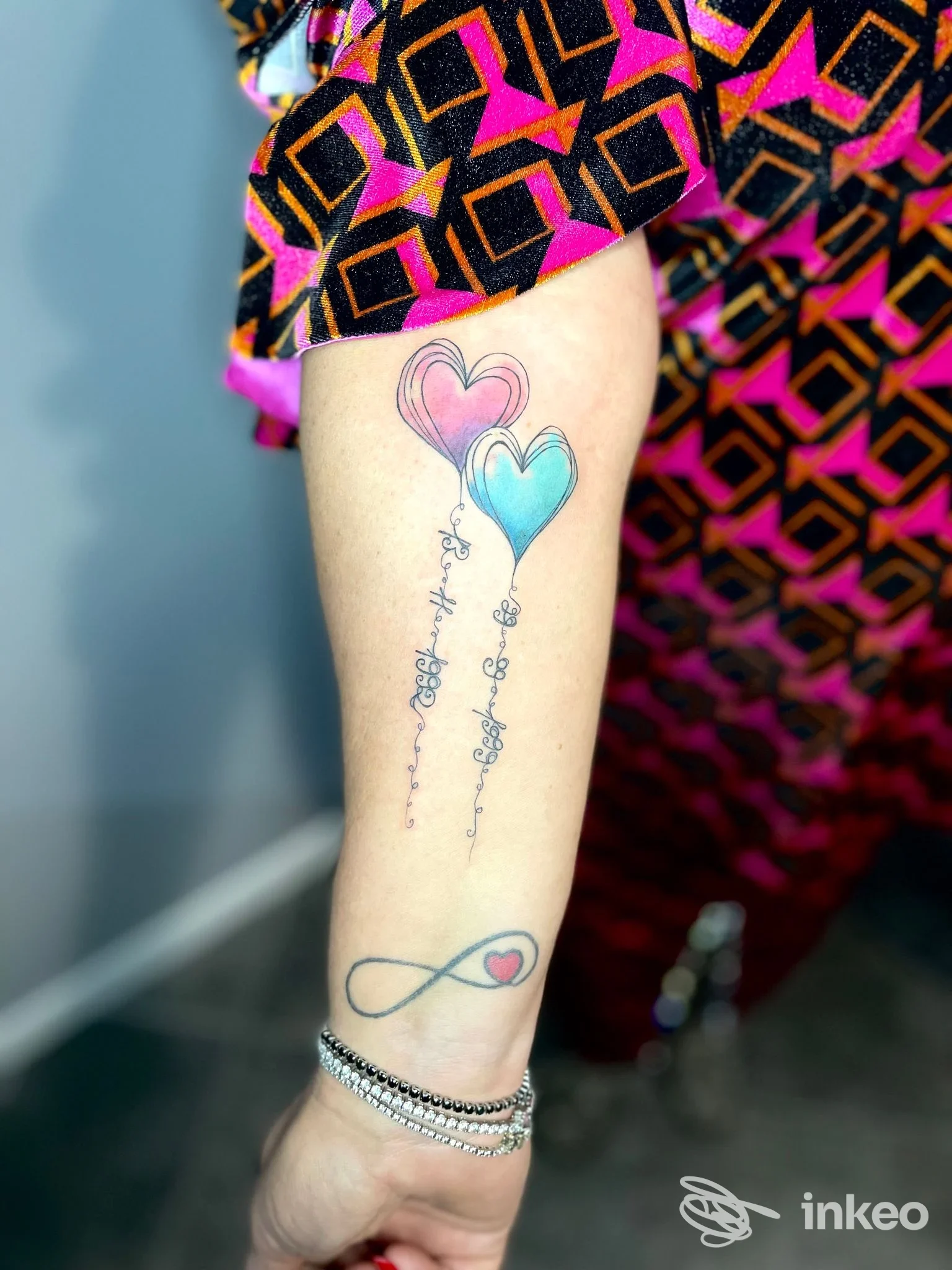 Tattoo su un avambraccio raffigurante due palloncini a cuore, rosa e azzurro, con una linea di testo e un infinito con cuoricino, visibile vicino al polso.
