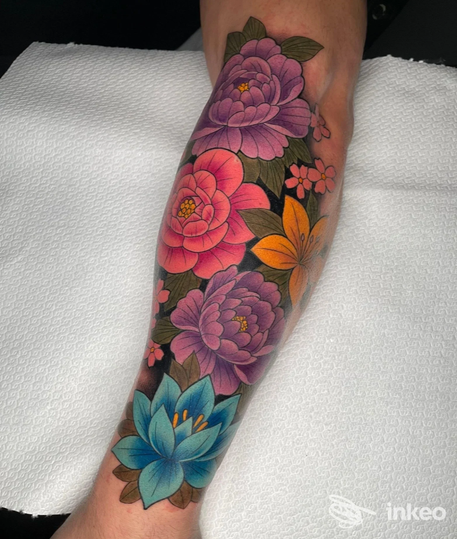 Tattoo floreale colorato su pelle visibile, grandi fiori rosa, viola e azzurro, realizzato in uno studio di tatuaggi. Parziale visibilità della gamba/artwork, senza volto.