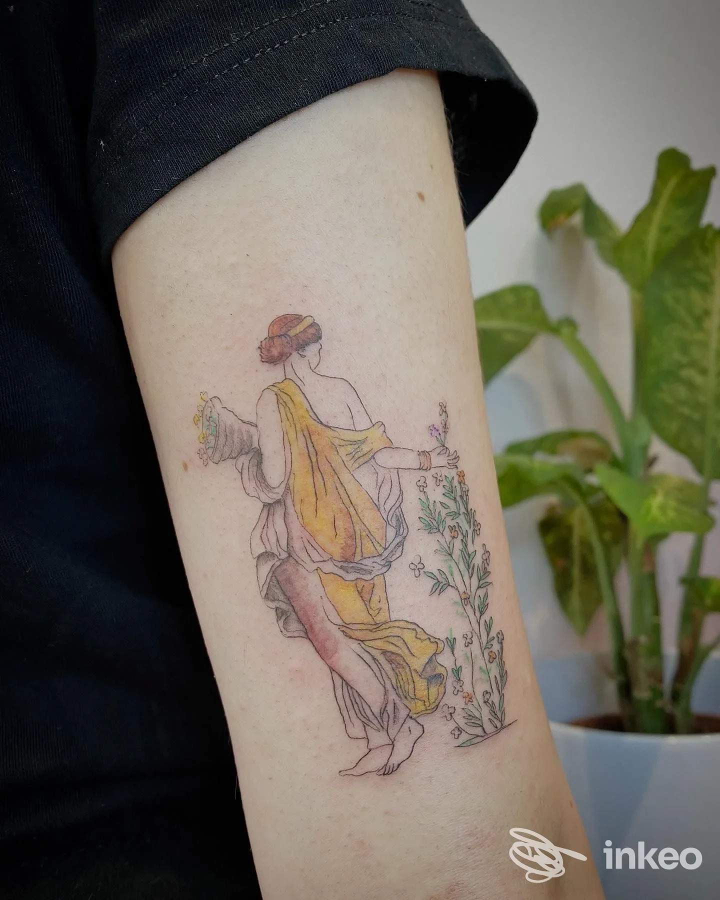 Tattoo colorato su avambraccio raffigurante una figura femminile con vestito giallo, contornata da piante, realizzato su pelle reale senza presenza di volto visibile.
