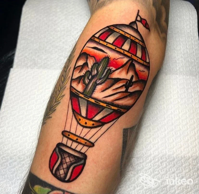 Tattoo colorato sull'avambraccio raffigurante una mongolfiera con paesaggio montano al tramonto, eseguito su pelle reale.