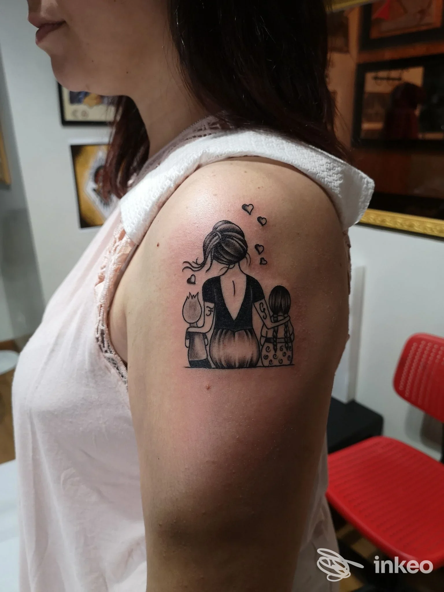 Tattoo sul braccio di una donna: una figura femminile abbraccia due bambini, contorni neri su pelle. cuori intorno e sfondo di sala tatuaggi.