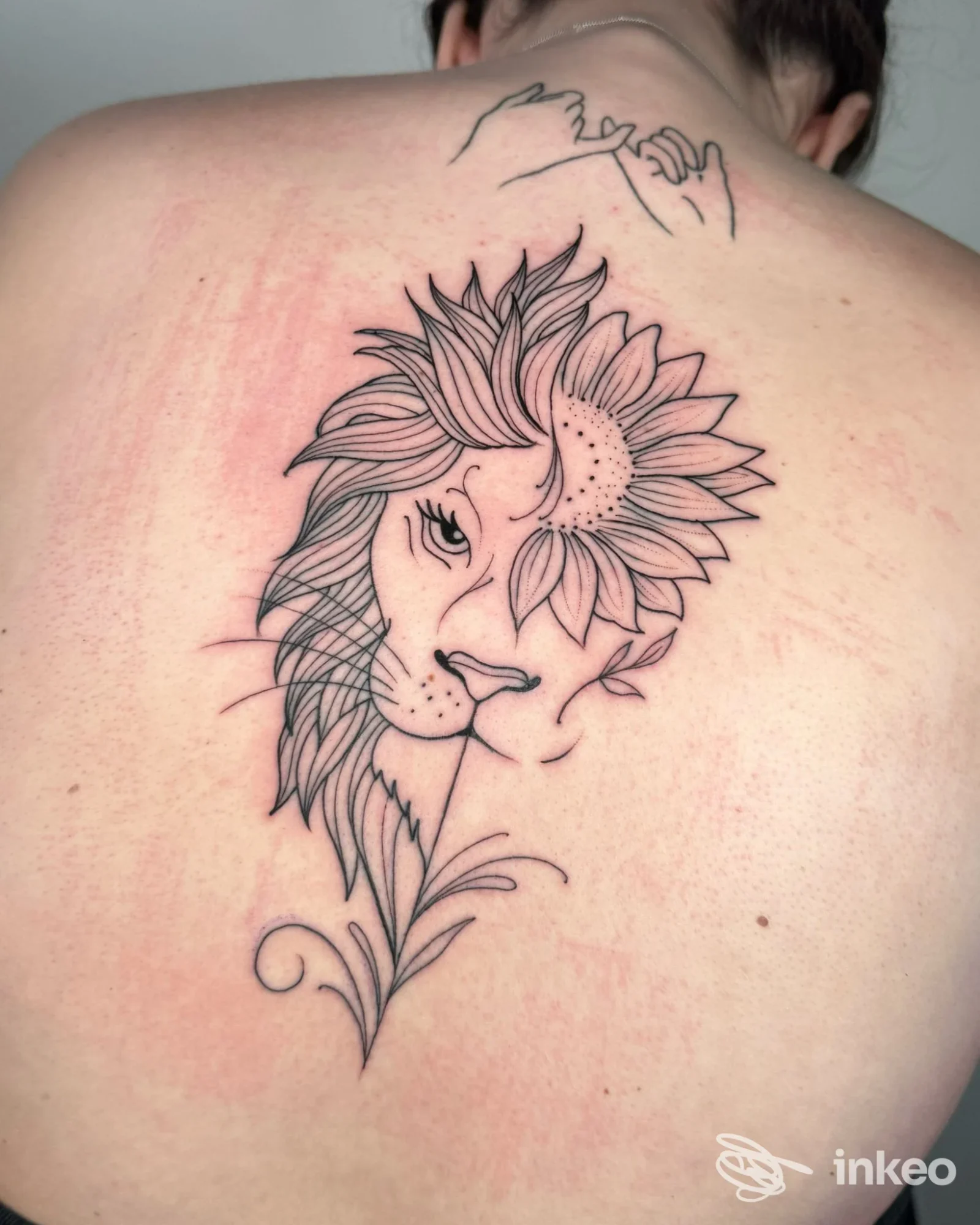 Tattoo visibile sulla spalla di una persona: ritratto stilizzato di leone con una grande girasola, linee nere pulite su pelle chiara, realizzato come tatuaggio permanente.