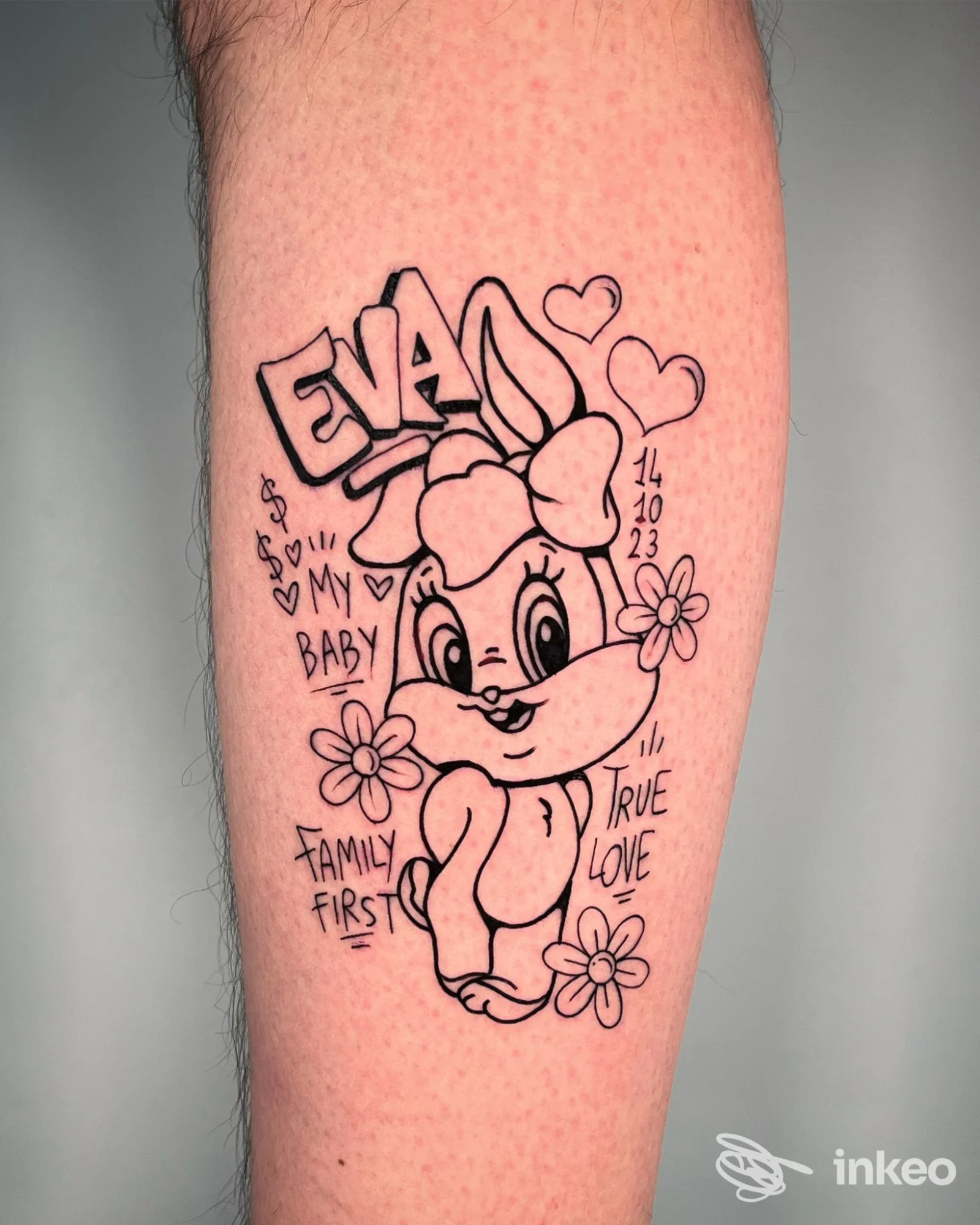 Braccio di una persona con un tatuaggio a contrasto nero che raffigura un coniglio cartone animato con fiori e cuori, testo e decorazioni intorno.