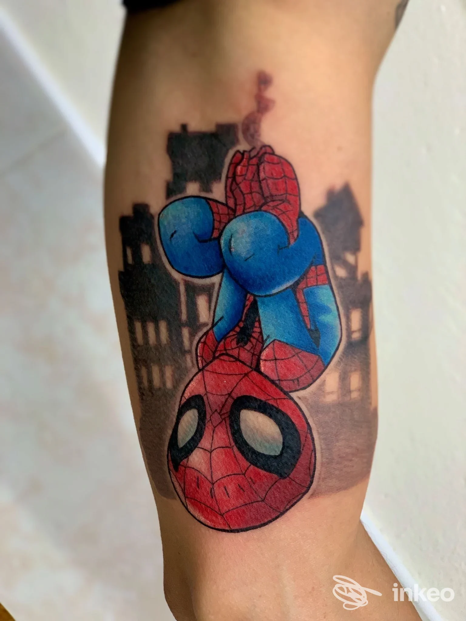 Tatuaggio colorato di Spider-Man capovolto sul braccio, con sfondo scuro a skyline e silhouette di edifici.