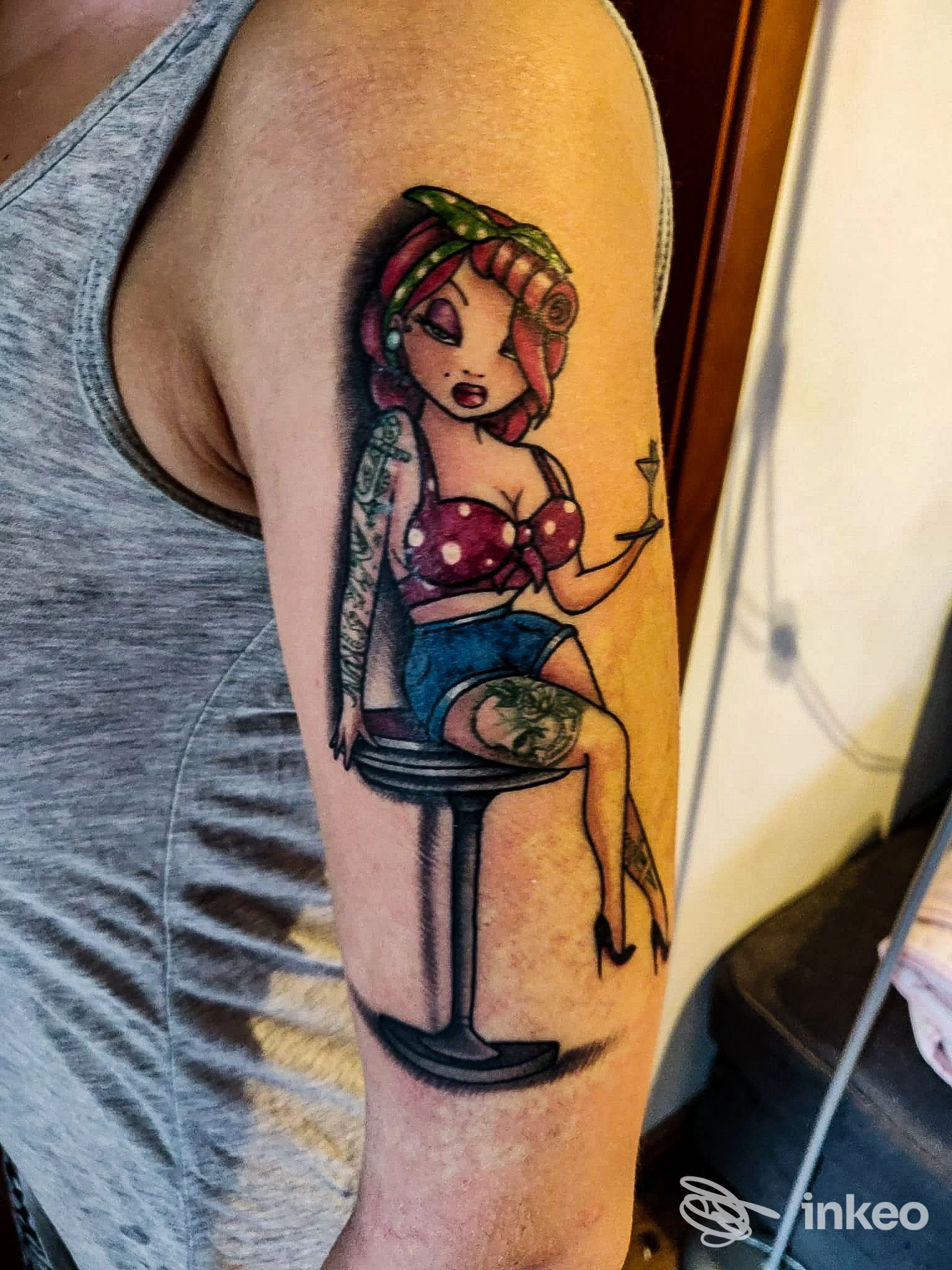 Tatuaggio su braccio raffigurante una ragazza in stile cartoon, seduta su uno sgabello, colori vivaci e dettagli a linea nera. L’immagine mostra la porzione superiore del braccio e la pelle è chiaramente tatuata.