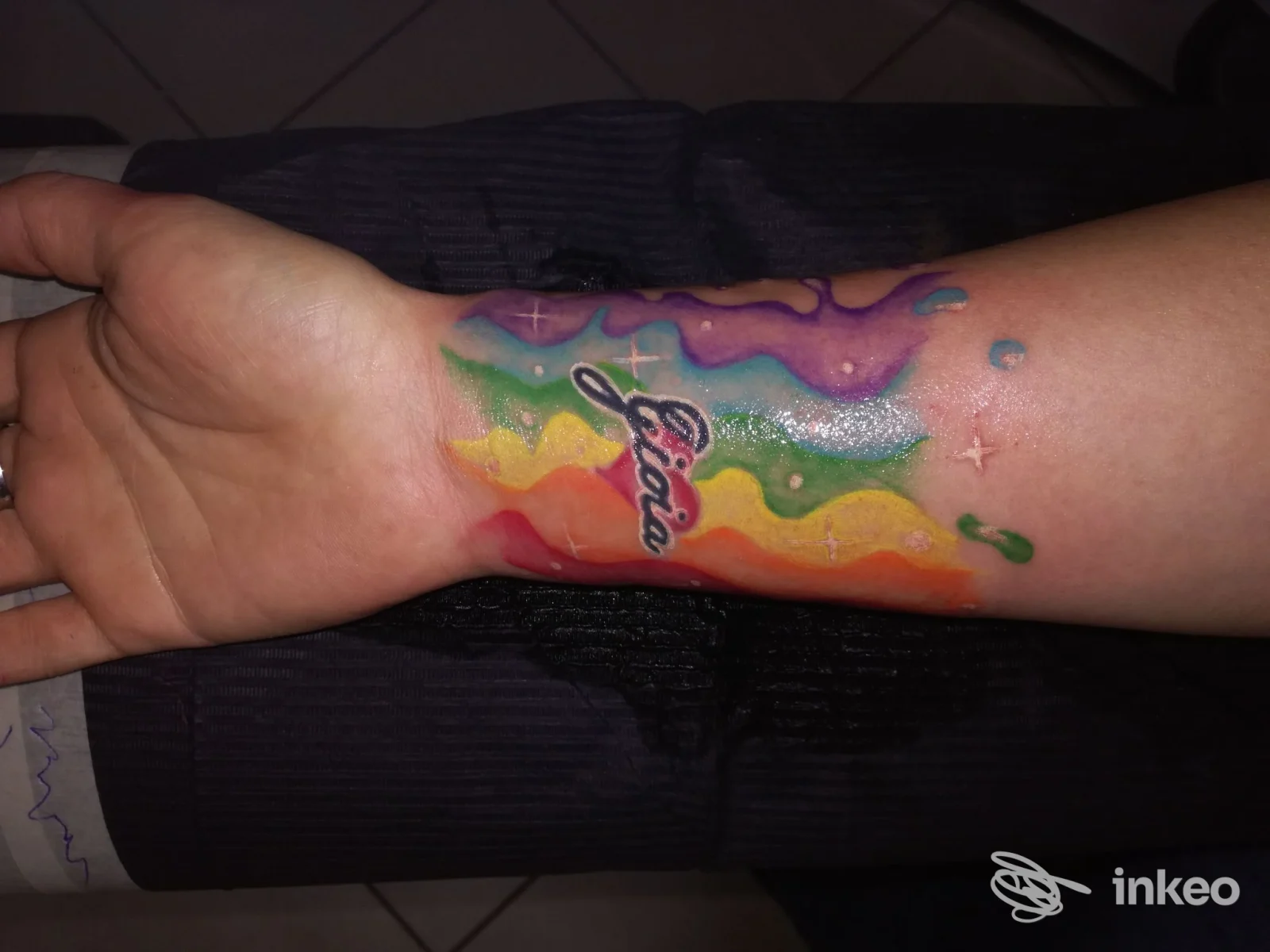 Avambraccio di una persona con un tatuaggio colorato a strisce arcobaleno e schizzi di vernice, testo in corsivo nero, realizzato da Mille Colori Tattoo a Cesano Maderno.
