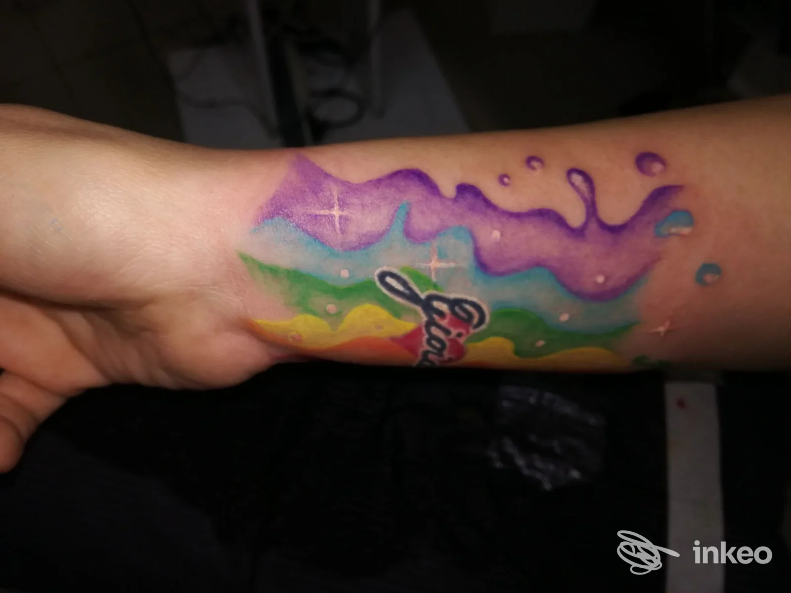 Avambraccio con tatuaggio colorato stile acquerello, sfumature viola, verde e giallo, testo in nero parzialmente visibile, senza volto umano inquadrato.