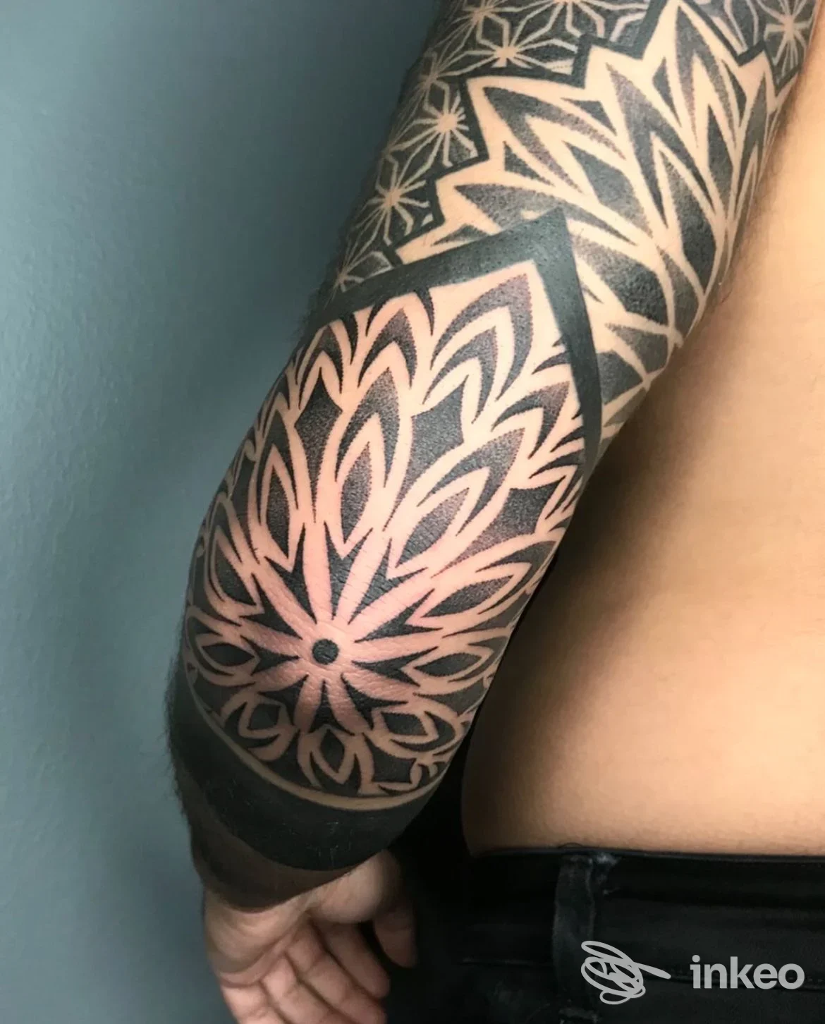 Tatuaggio geometrico a mandala su braccio, disegno nero su pelle chiara, bordo del braccio visibile, nessun volto presente.