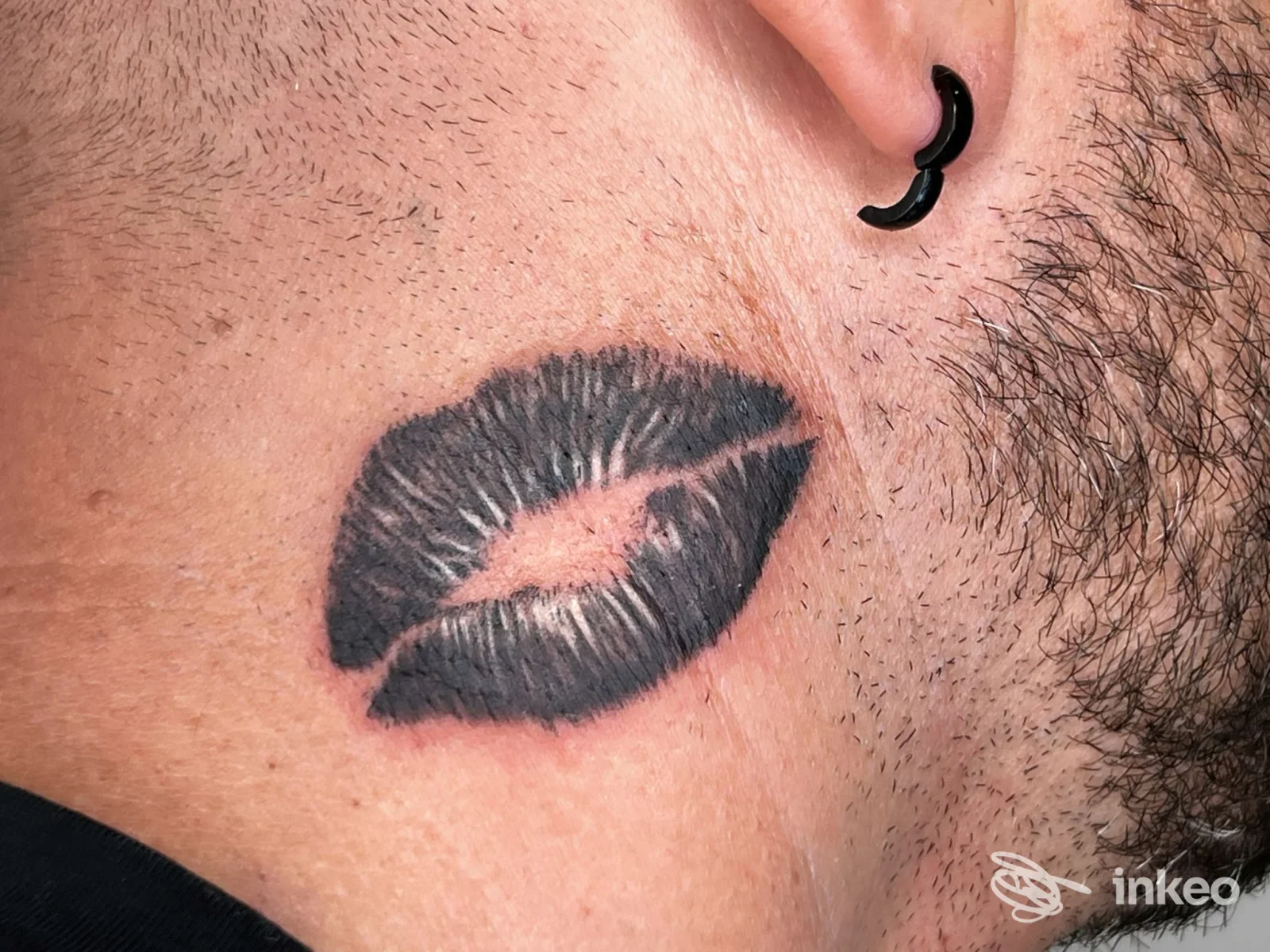 Tattoo a labbra scure su pelle reale dietro l’orecchio, vicino al collo; orecchino nero visibile.