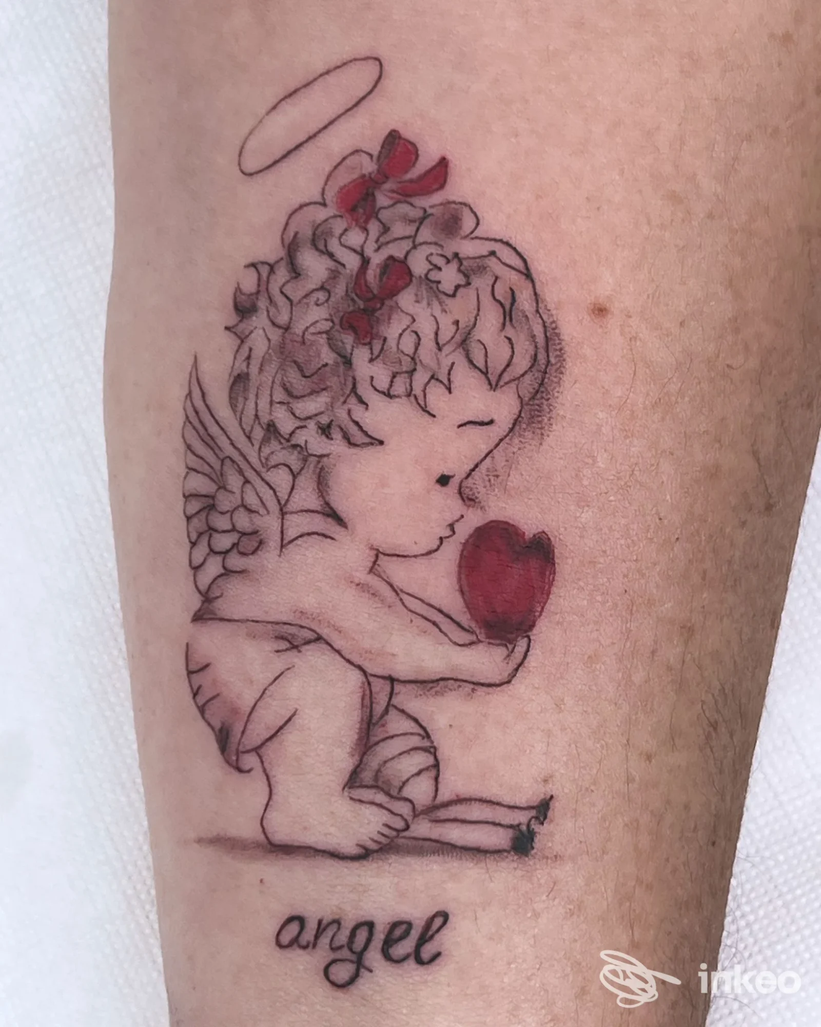 Tattoo su pelle di un angelo bambino alato che tiene un cuore rosso, con linee nette e sfumature, visibile sullavambraccio; nessun overlay sociale. Studio Antonella Nail Tatoo.