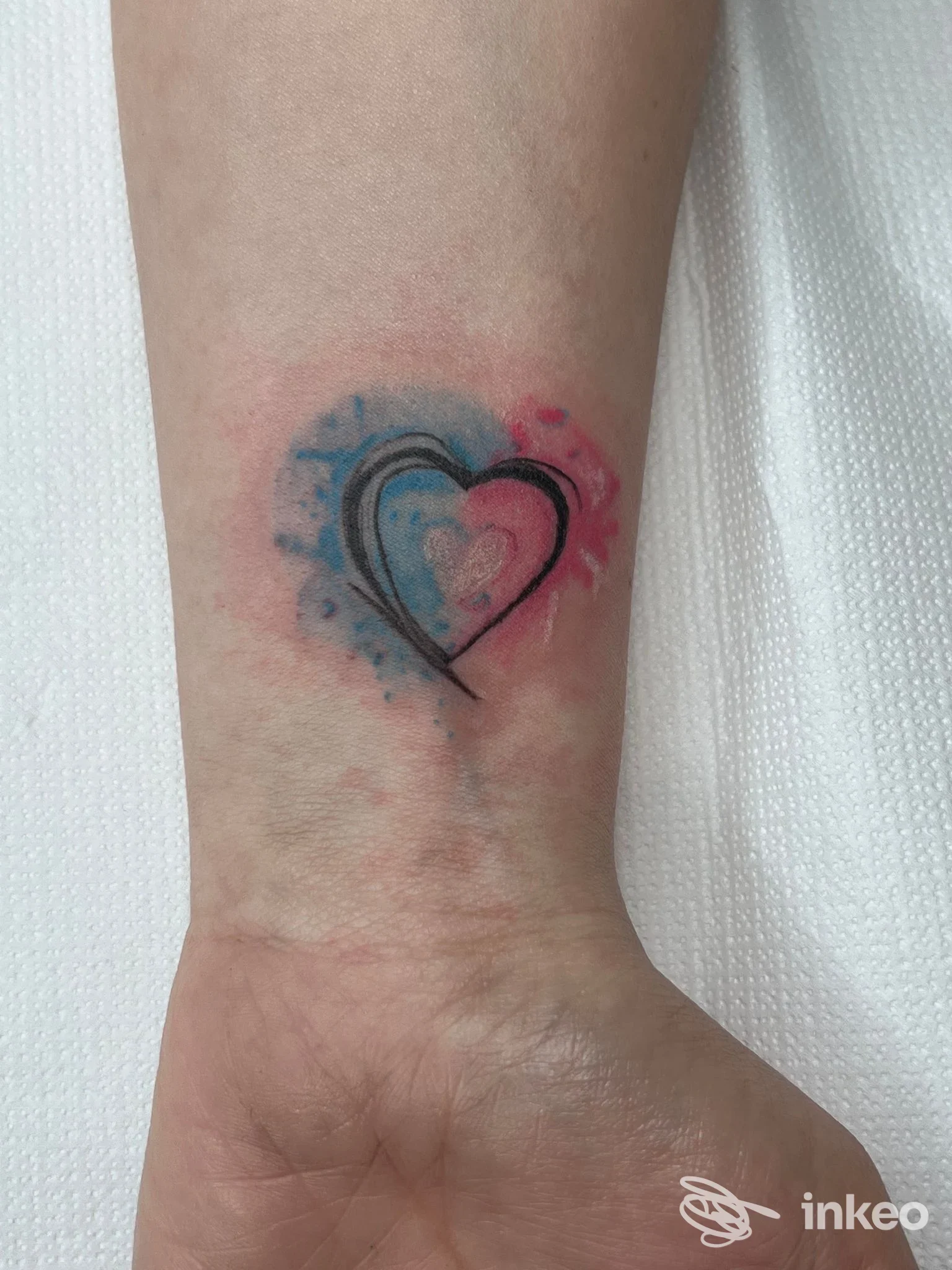 Piccolo tatuaggio sul polso: cuore disegnato con contorni neri, bordi sfumati da tonalità blu e rosa, su pelle chiara.
