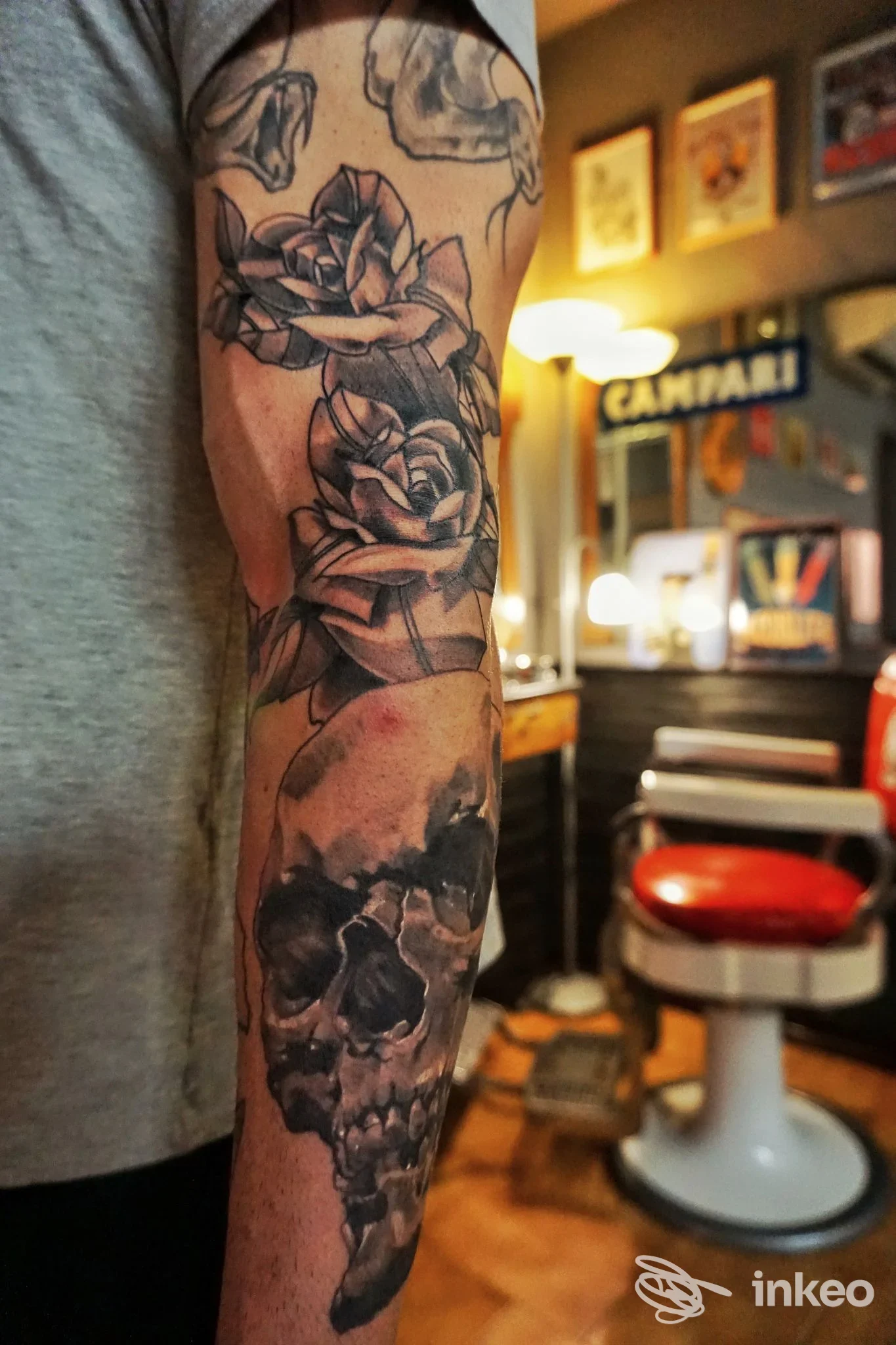 Braccio sinistro con una manica di tatuaggi che ritrae rose e un teschio in tonalità scure, sullo sfondo un salone.