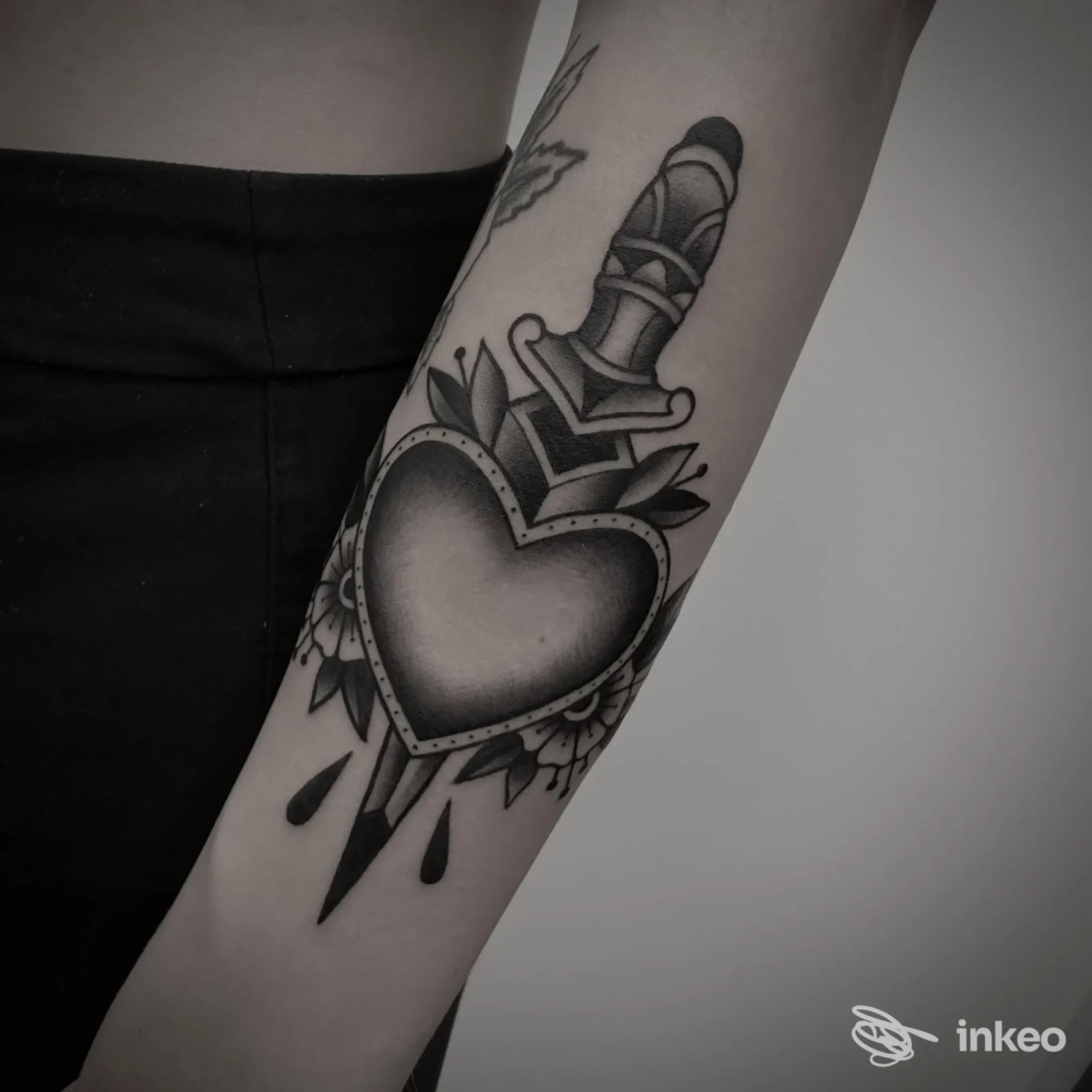 Peltro tono grigio, un tattoo sul braccio raffigura un cuore con daga, ornamenti floreali e gocce, in bianco e nero, visibile in primo piano su sfondo neutro.