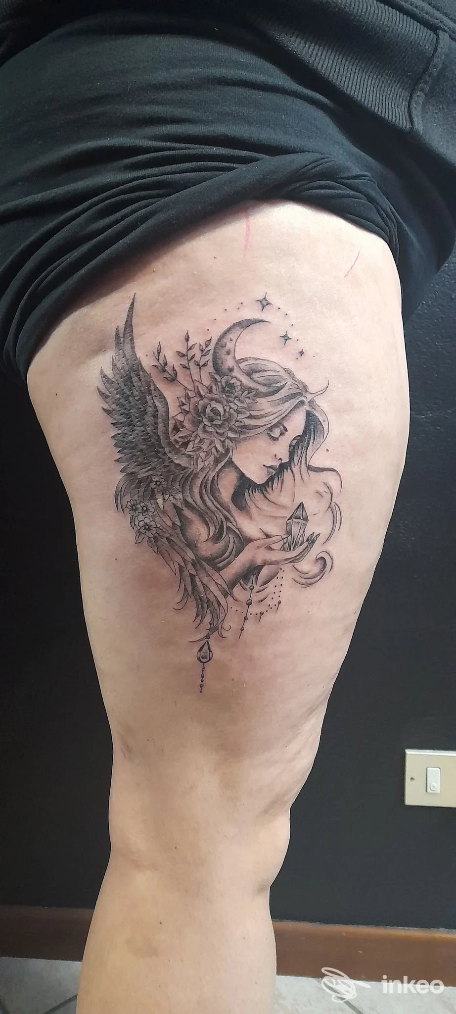 Tattoo nero su pelle raffigurante una figura alata con luna e stelle, situata sulla coscia di una persona, con dettagli finemente lavorati.