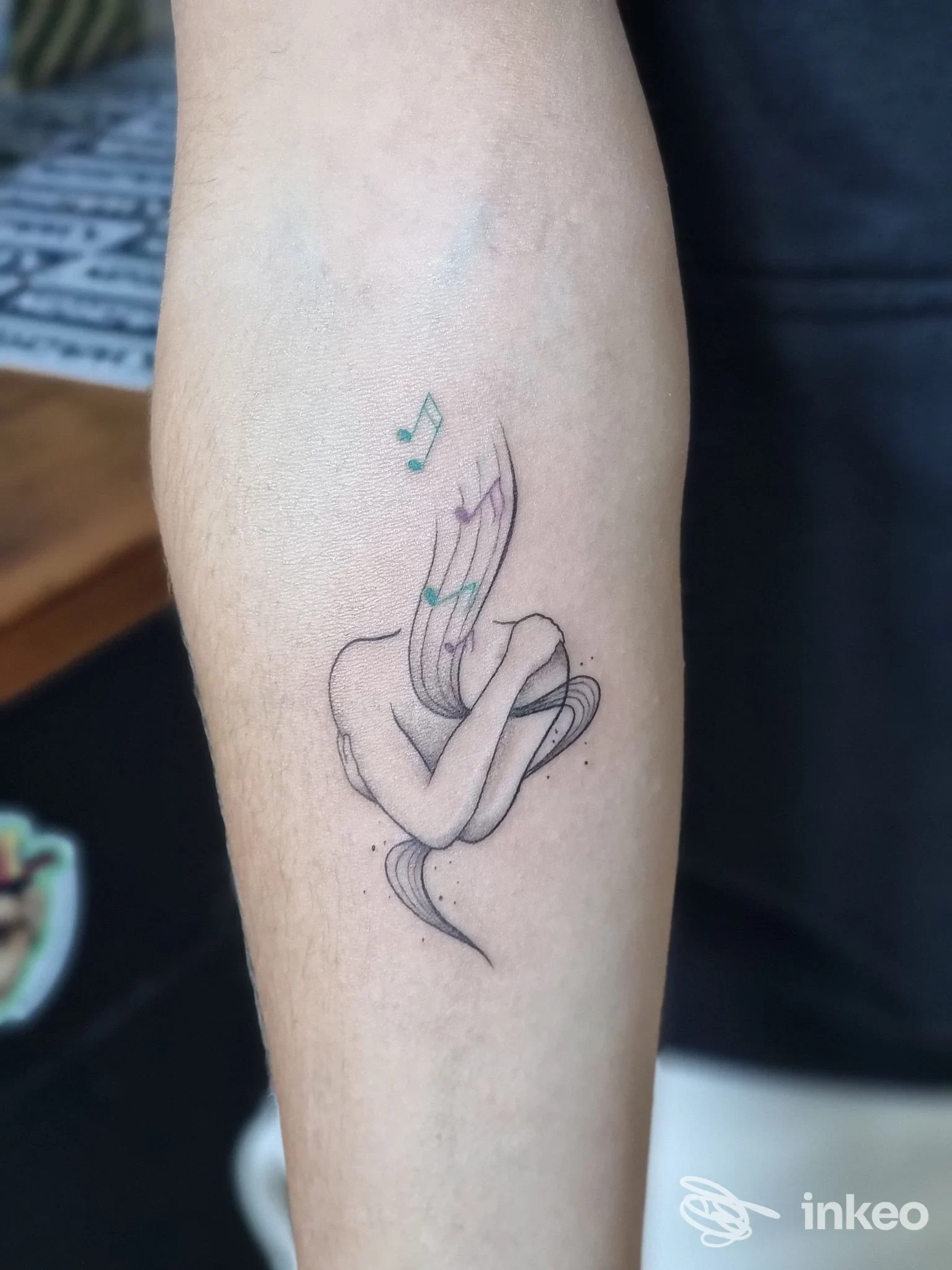 Tattoo su avambraccio raffigurante una figura stilizzata che abbraccia le ginocchia, con note musicali colorate sopra.