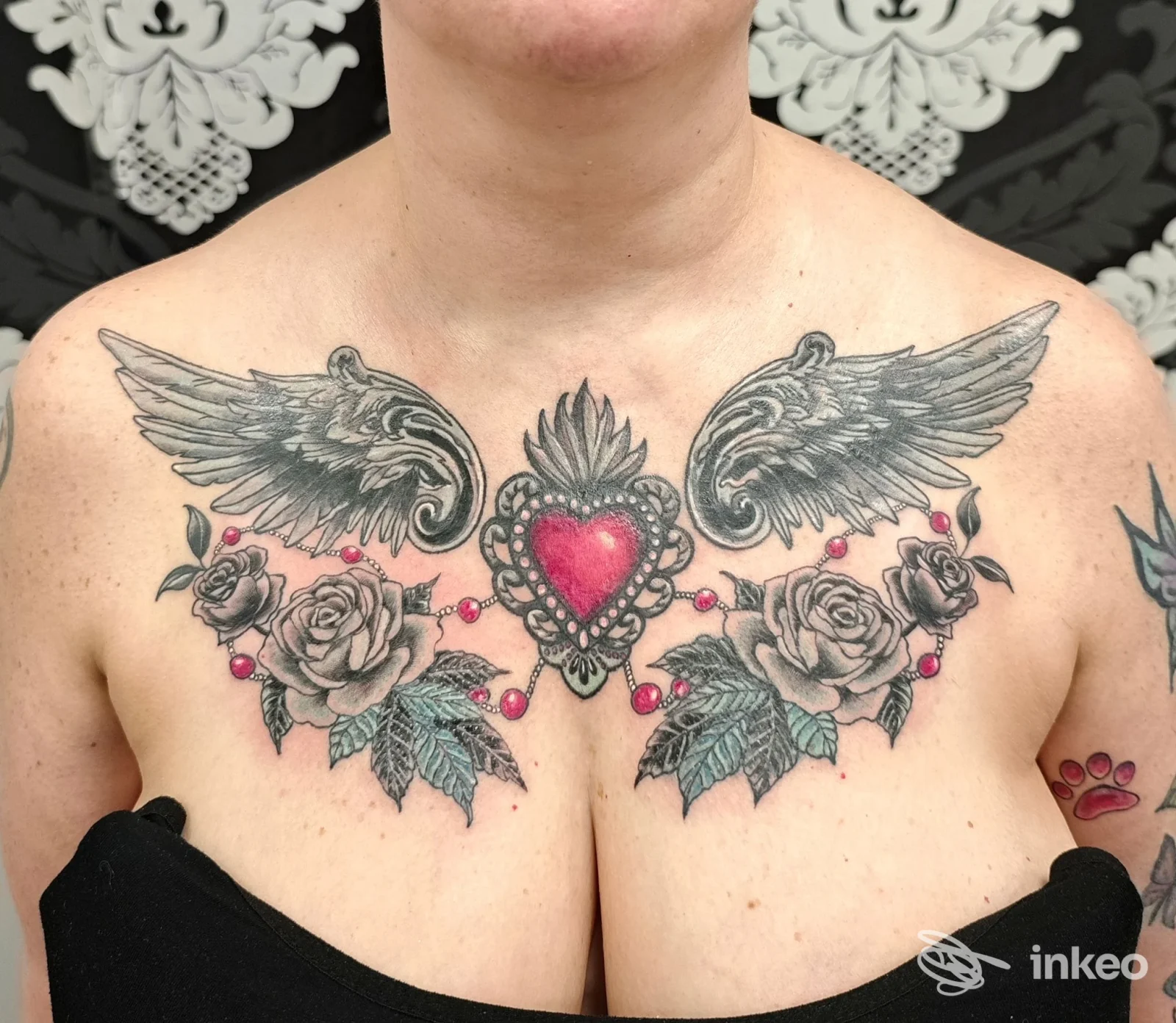 Tattoo sul petto con ali ai lati e cuore centrale, accompagnato da rose decorative; visibile parte del collo, nessun overlay o testo promozionale, foto in alta risoluzione.