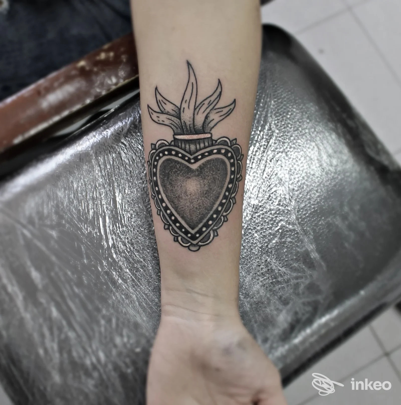 Tatuaggio a cuore con decorazioni e fiamme sulla pelle dell’avambraccio, eseguito in stile linework, senza presenza del viso.