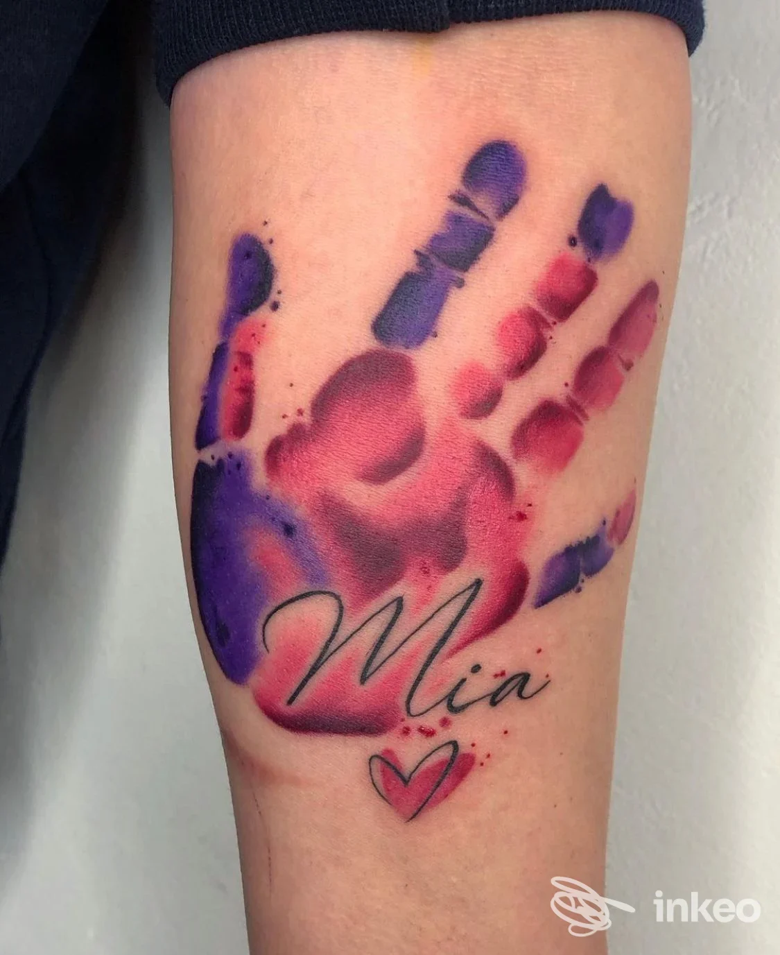 Foto di un braccio con un tatuaggio colorato a impronta di mano viola e rosso, con scritto Mia e un piccolo cuore.