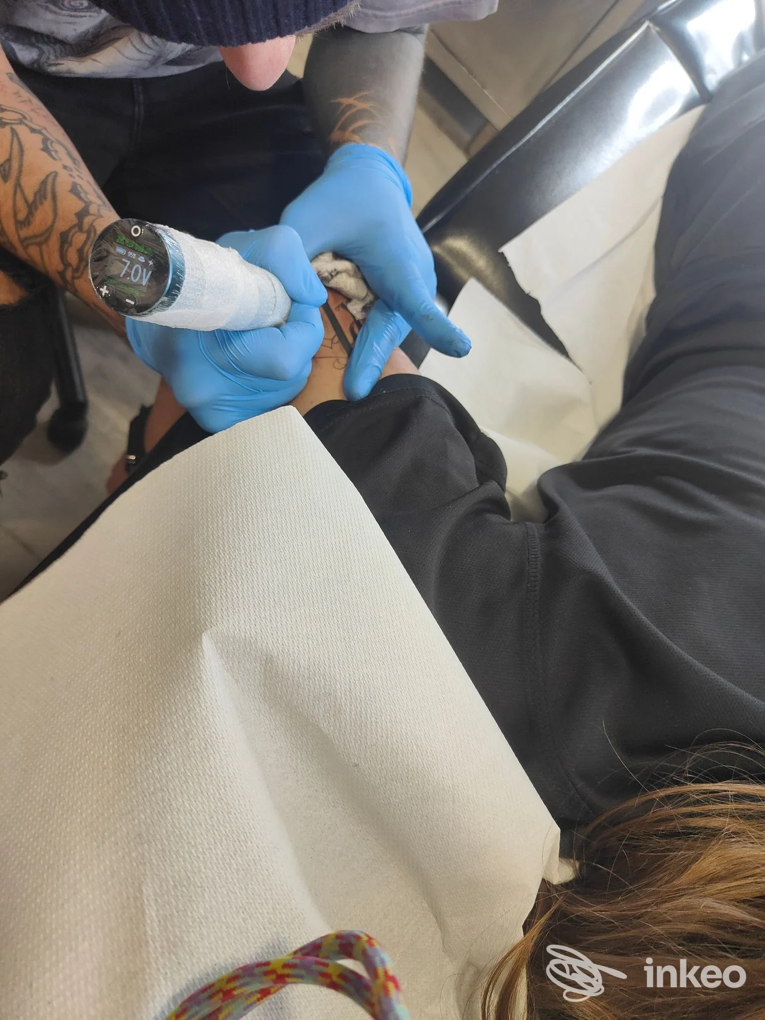 Primo piano di una sessione di tatuaggio sull'avambraccio: una mano con guanti blu maneggia una macchina da tatuaggi sulla pelle, nessun volto visibile.