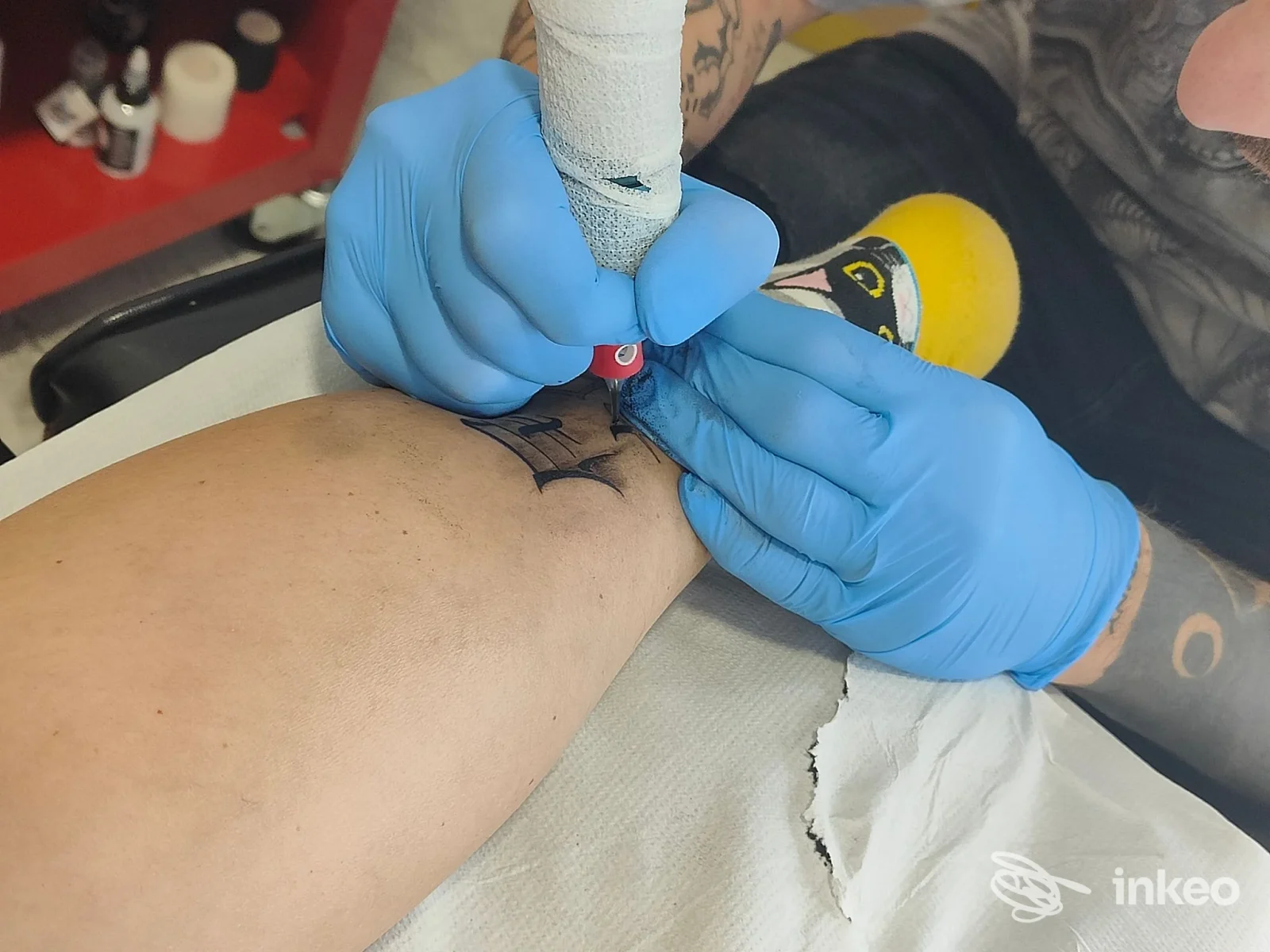 Primo piano di un tatuaggio in corso sull’avambraccio, con guanti blu che lavorano sulla pelle in uno studio chiamato Rocket Tattoo Lab.