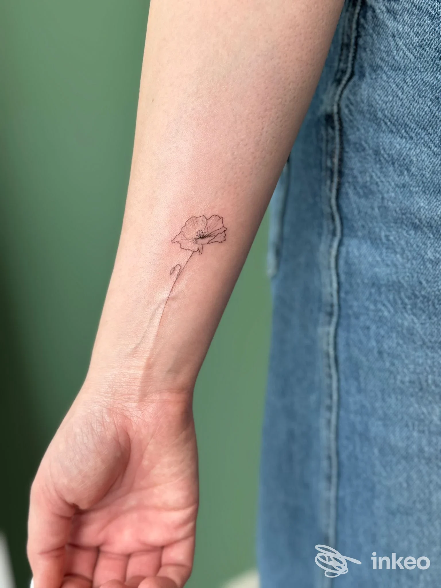Piccola tatuatura di un fiore su avambraccio sinistro, realistica e ben definita, scattata all’aperto vicino a una maglietta in jeans. Studio RunawayLab, Brescia.