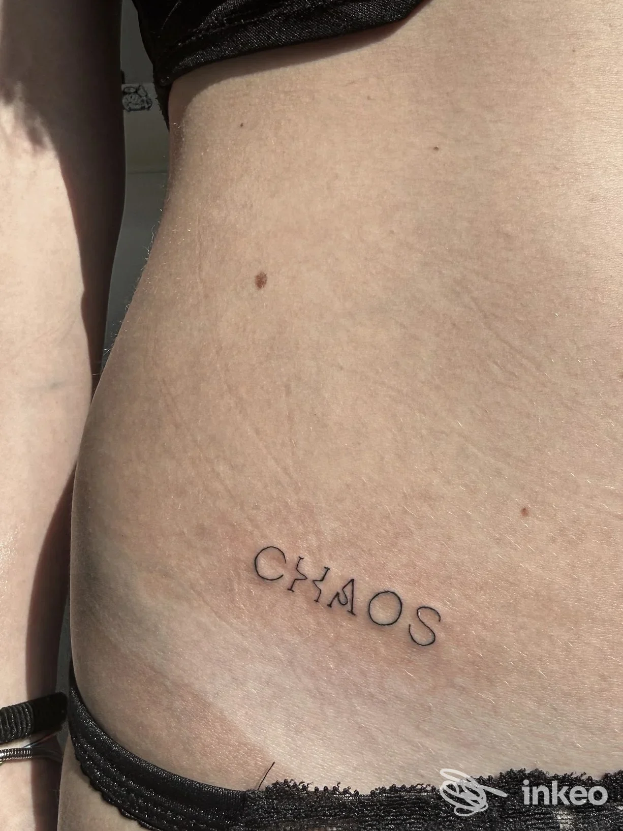 Tattoo nero con la parola CHAOS sul fianco sinistro, visibile sulla pelle, parte laterale del torso.