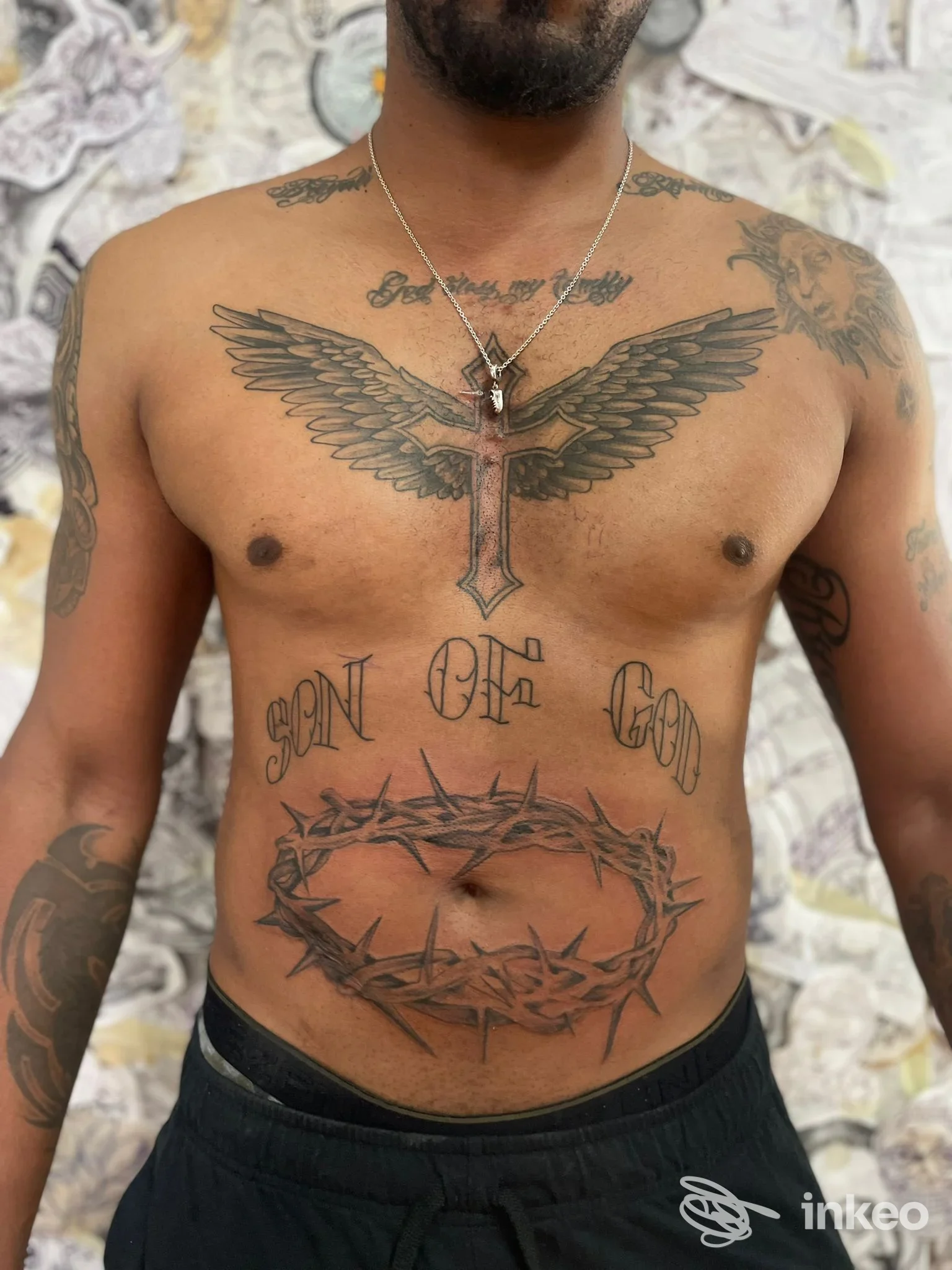 Uomo senza maglietta con grandi tatuaggi sul torace: ali stellate sul petto, scritte sul busto, corona di spine sull’addome, girocollo con ciondolo in primo piano.