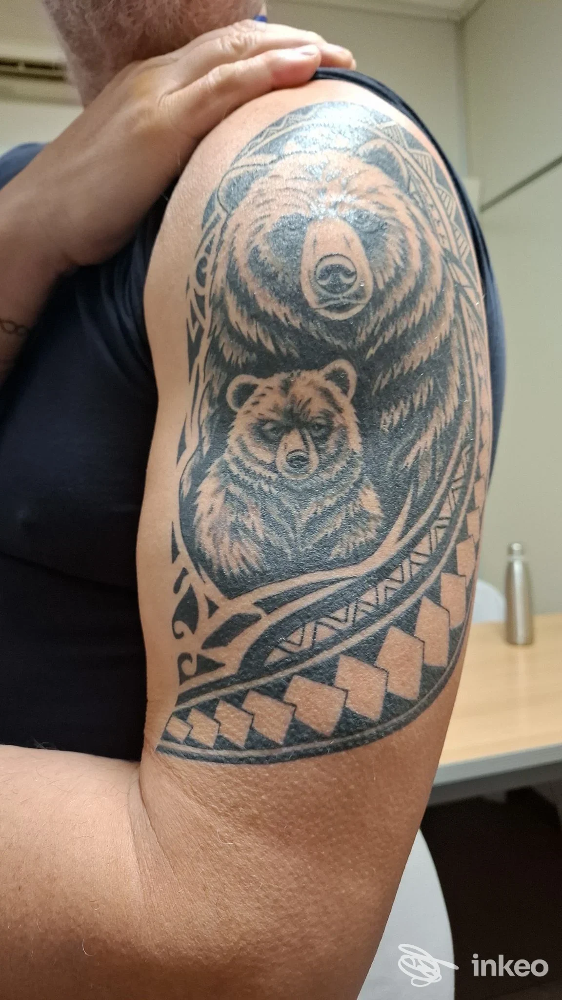 Tatuaggio realistico sul braccio con due orsi, adulto e cucciolo, racchiusi in una cornice ornamentale e un motivo a scacchi lungo il bordo inferiore.