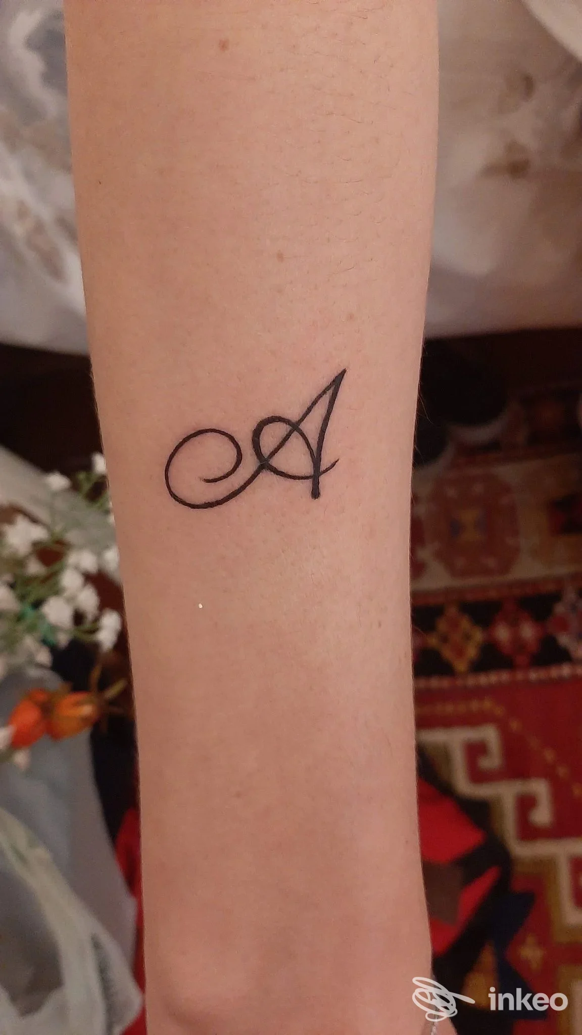 Tattoo singolo sull'avambraccio: lettera corsiva 'A' nera, realizzazione corporea visibile, nessun overlay sociale.