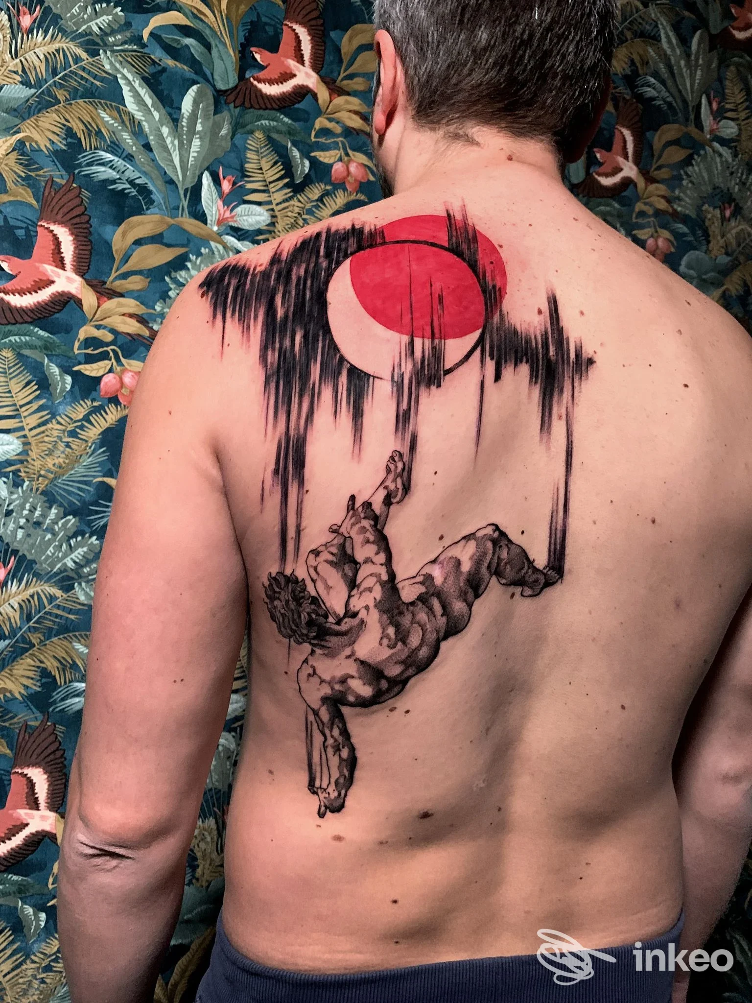 Tattoo sul dorso di un uomo: una figura pendente si aggrappa a una linea nera con un sole rosso sopra, su sfondo di carta da parati floreale. Vestito poco sotto, è visto di schiena.