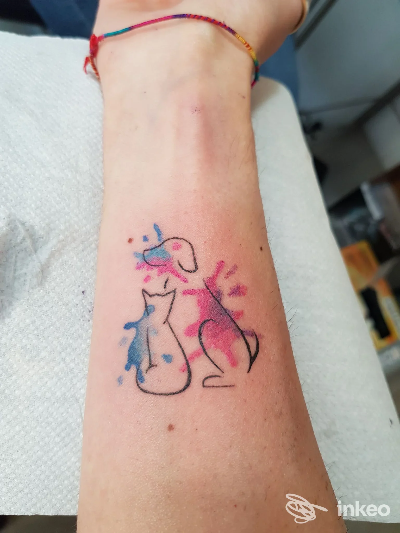Piccolo tatuaggio di due gatti stilizzati con schizzi colorati a acquerello sul polso: linea nera con tocchi rosa e blu.