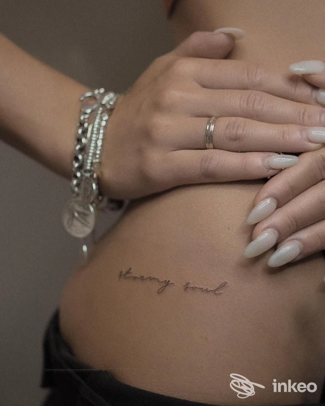 Tatuaggio testuale visibile sul fianco sinistro di una persona, con mani e bracciali in primo piano; nessun volto interamente visibile.