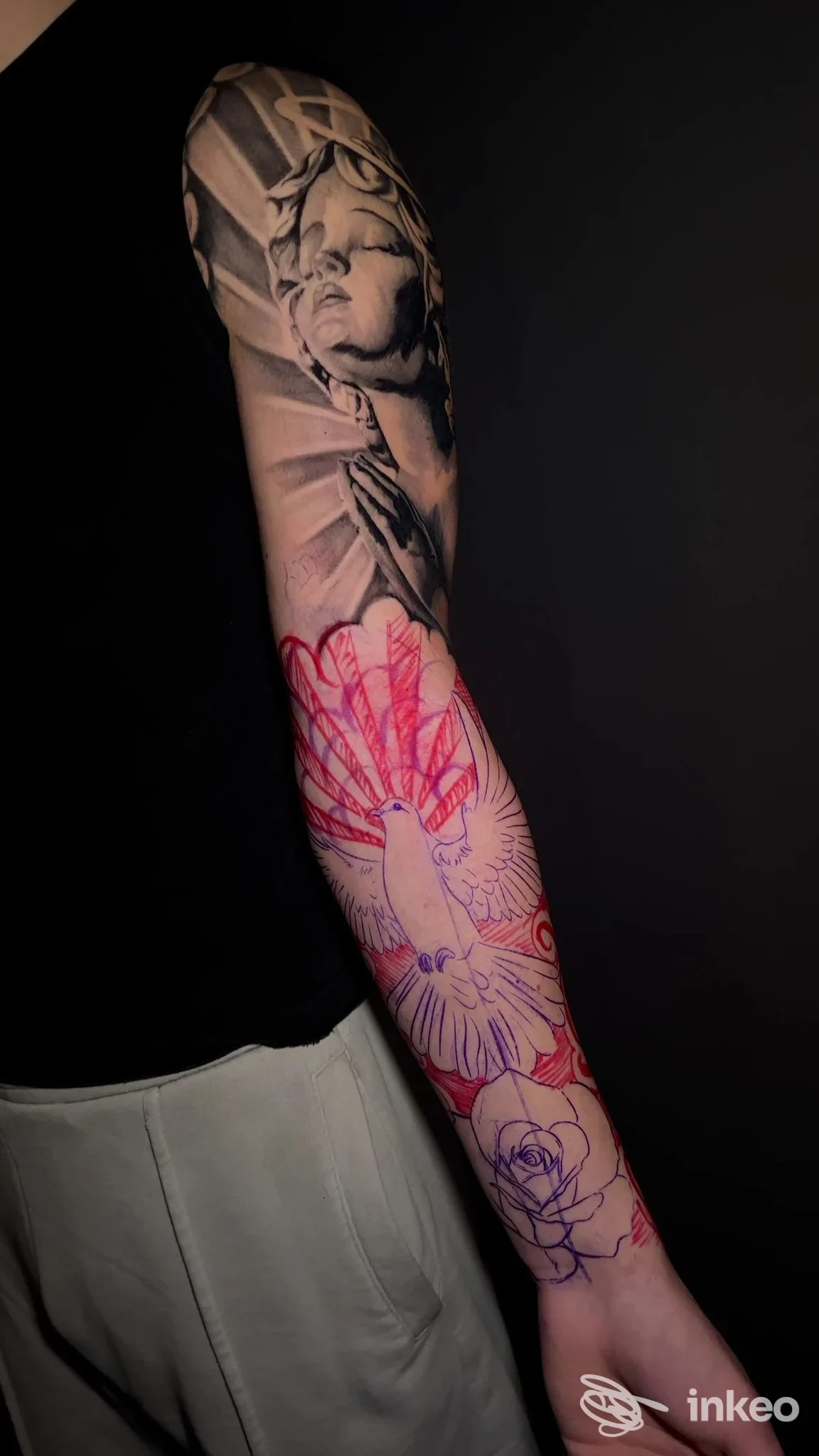 Braccio sinistro di una persona visto di profilo, tattoo reale sulla pelle: dettaglio grigio sul braccio superiore con figura, e sul polso una parte colorata rossa/rosa con schizzi floreali.