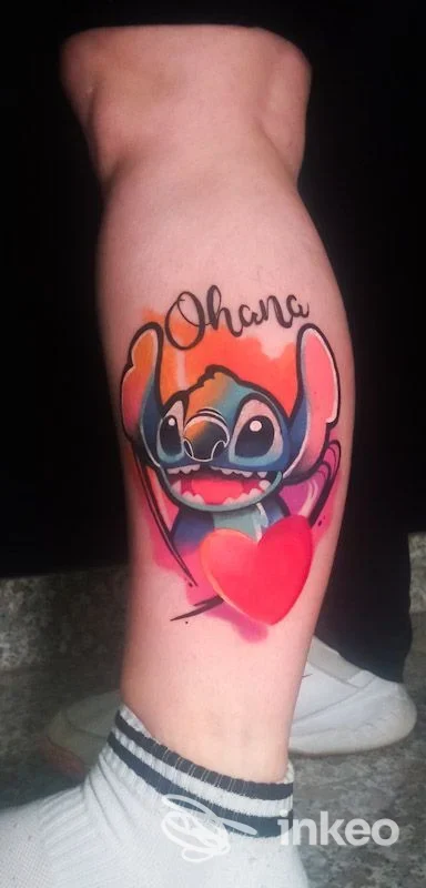 Tattoo a colori di Stitch che tiene un cuore rosso, situato sulla gamba sinistra, visibile chiaramente su pelle. Nessun volto umano visibile. Studio Apocalypse Tattoo, città non specificata.