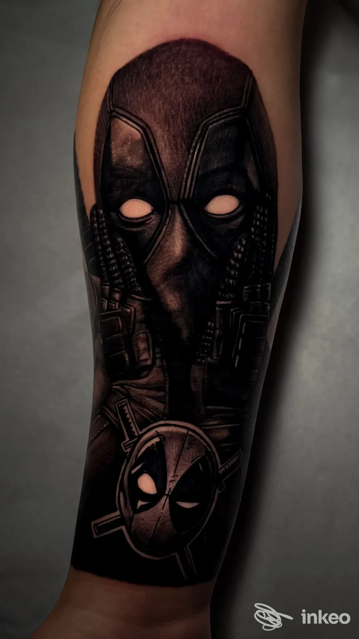 Tattoo realistico sul braccio raffigurante Deadpool con maschera, accompagnato da elementi di armi lungo l’avambraccio; esecuzione scura e dettagliata.
