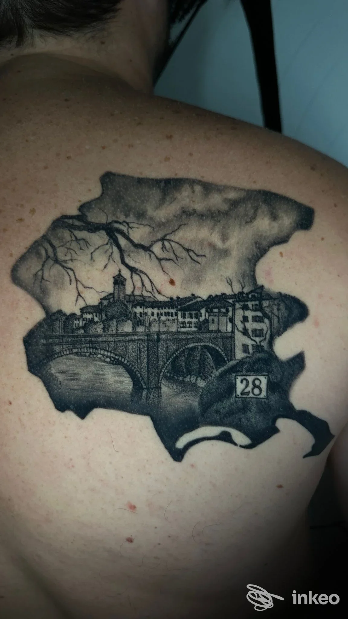Tattoo in bianco e nero sulla parte alta della schiena, raffigura una scena urbana con ponte all’interno di una forma irregolare simile a una mappa.