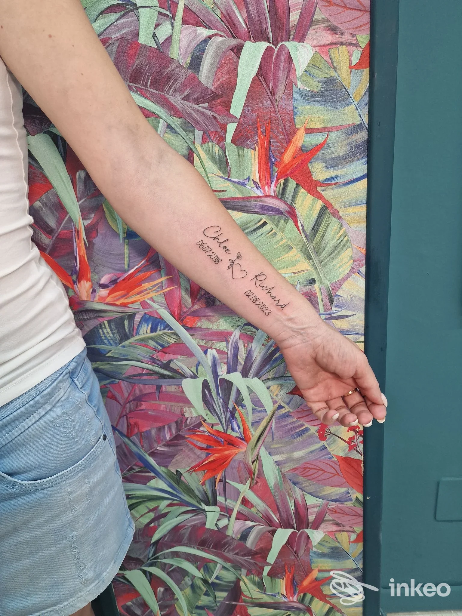 Avambraccio di una persona con un testo tatuato in corsivo sulla pelle, sfondo floreale colorato.