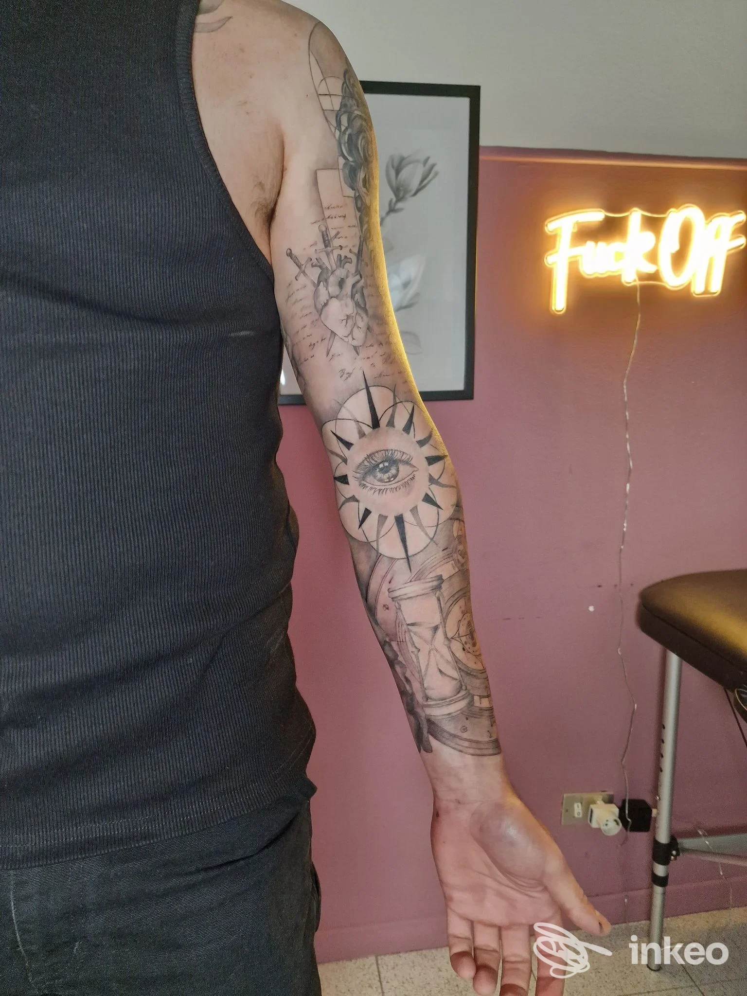 Braccio di una persona con tatuaggio visibile: design con occhio centrale incorniciato da elementi geometrici su pelle chiara, sfondo rosa con pannelli e neon.