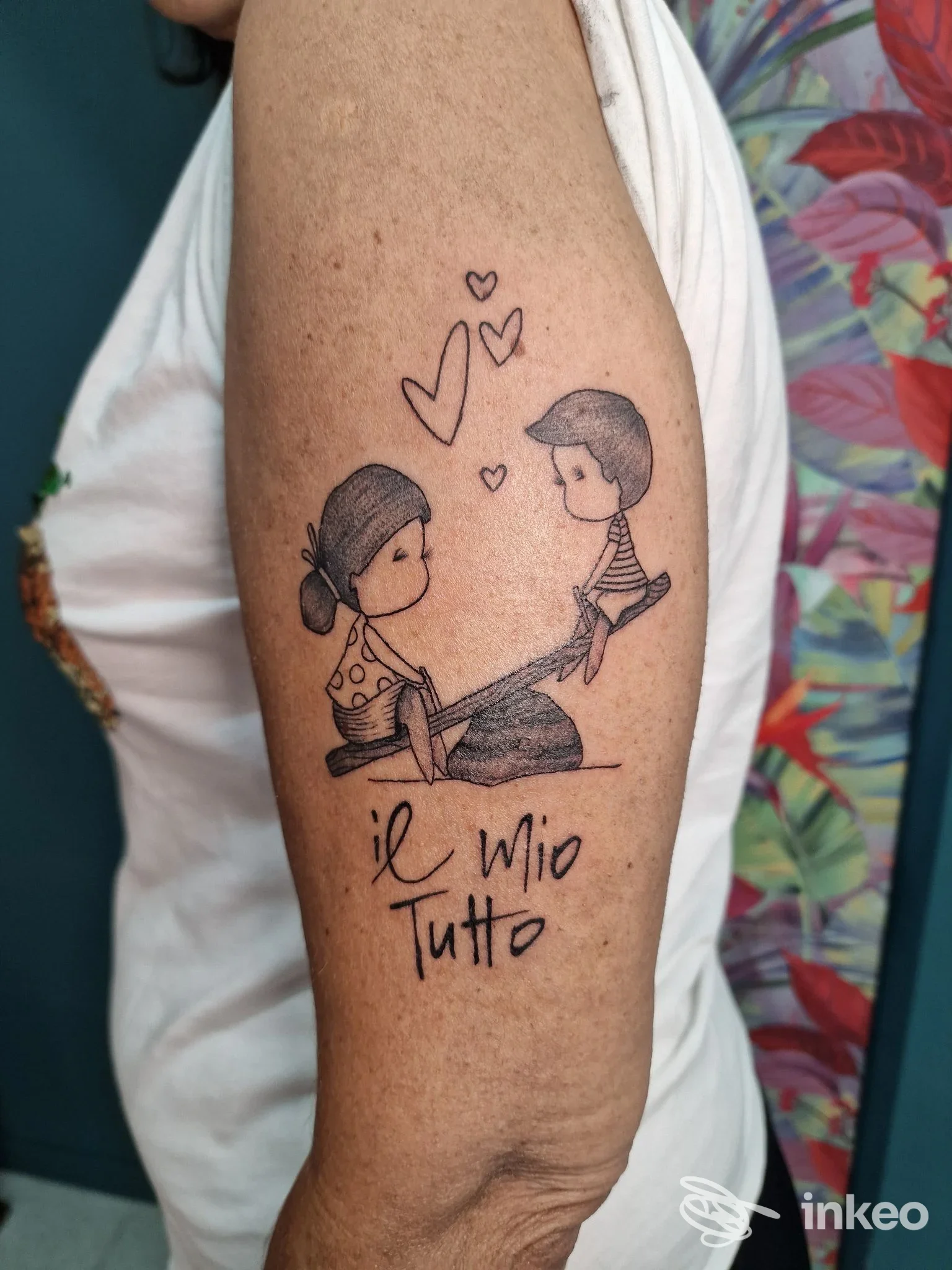 Tatuaggio visibile su braccio raffigurante due bambini che si prendono per mano con cuori tra loro e testo Il Mio Tutto, realizzato sulla pelle.