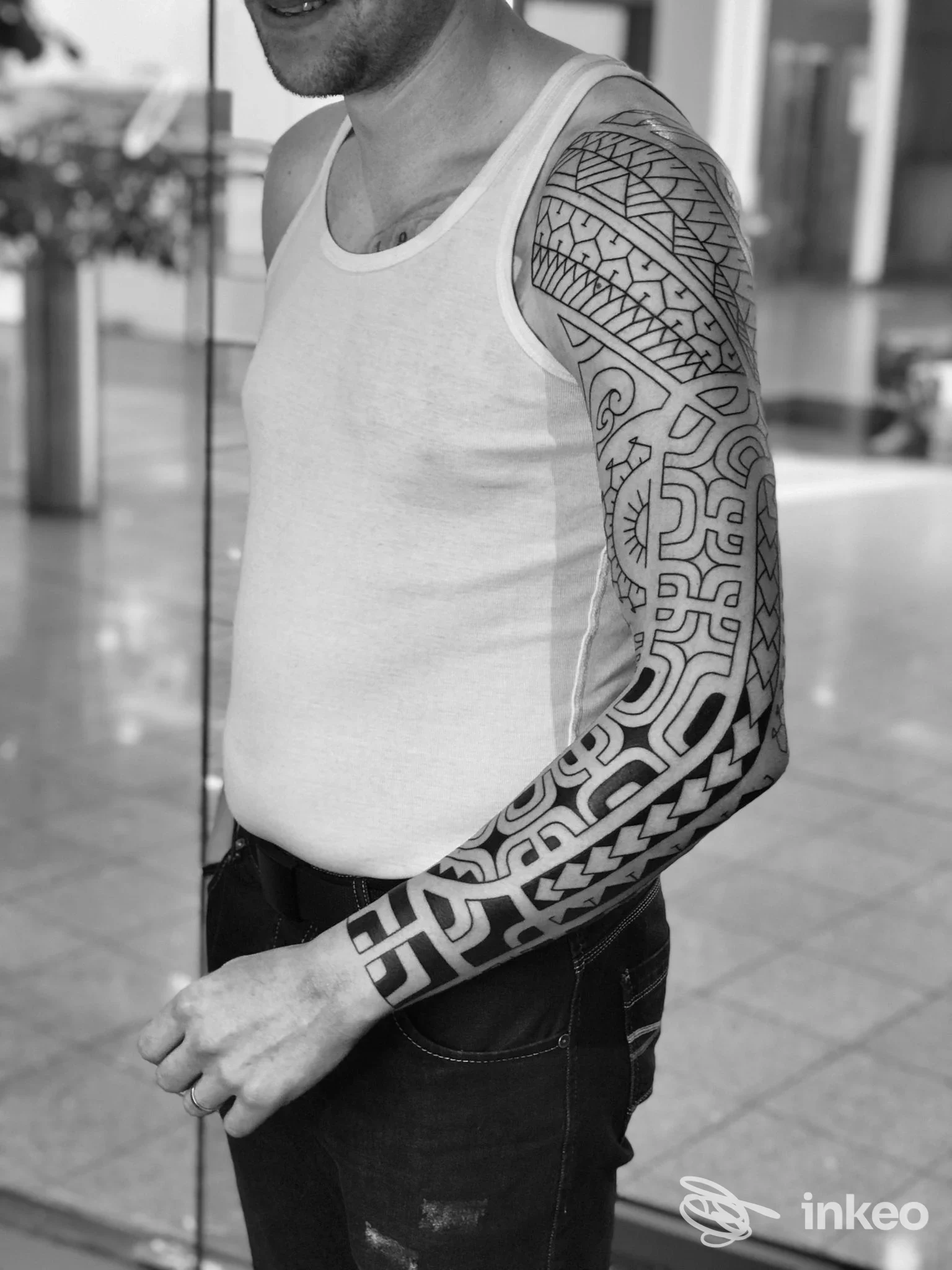 Uomo in canotta mostra un braccio sinistro completamente tatuato con motivi geometrici neri in stile blackwork. Foto ambientata in uno spazio urbano.