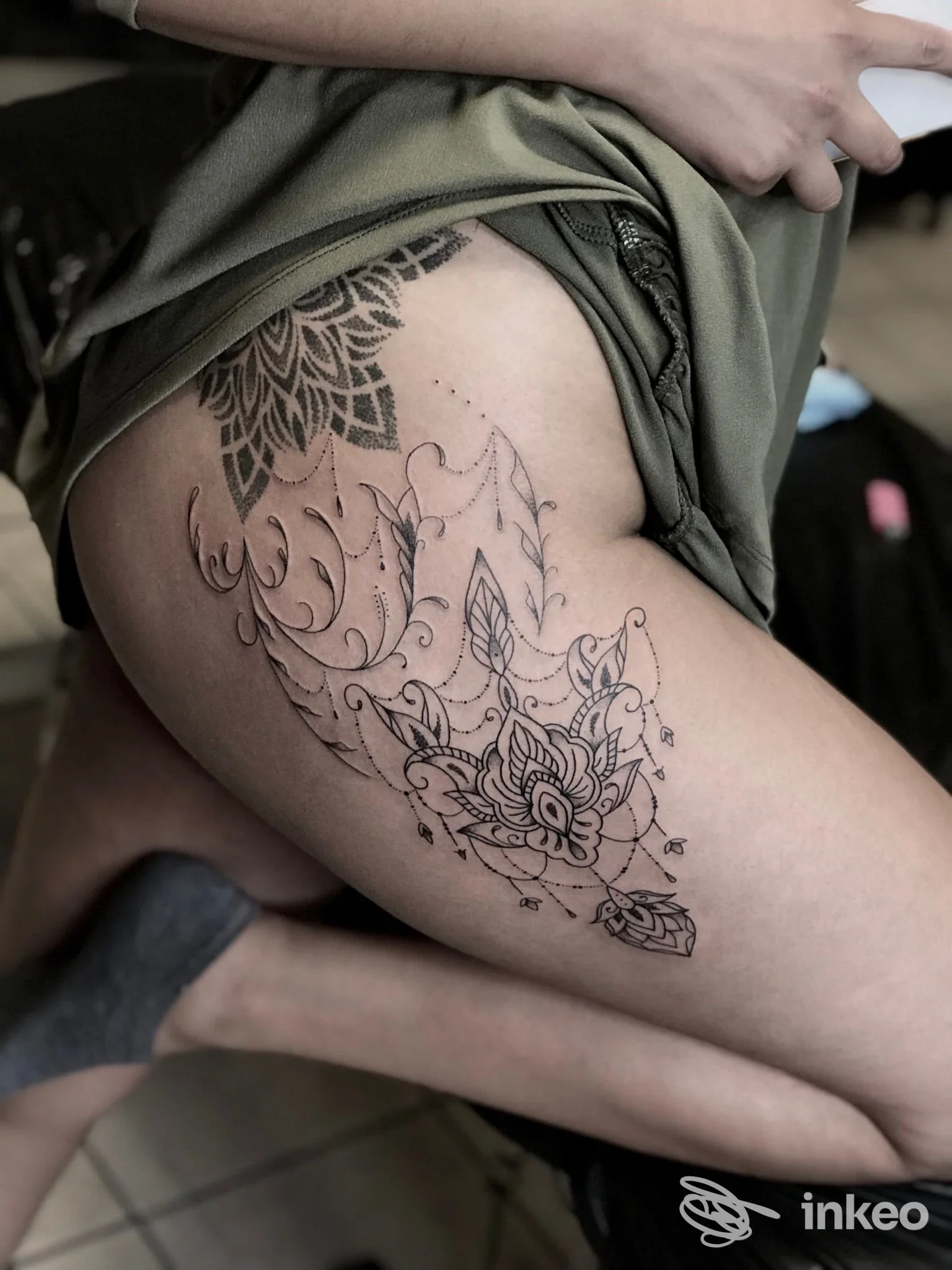 Tattoo floreale stilizzato sulla coscia esterna di una persona, con linee nere nette e riempimenti, visibile durante un abbassamento di tessuto. Studio Kiavash Tattoo.