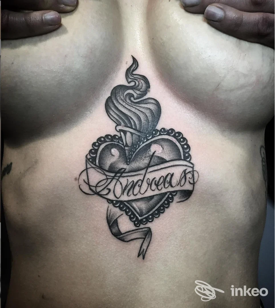 Tattoo realizzata sul torace: cuore con bordi decorativi e una ribbon banner con nome inciso, realistica in toni neri e grigi, visibile su pelle.