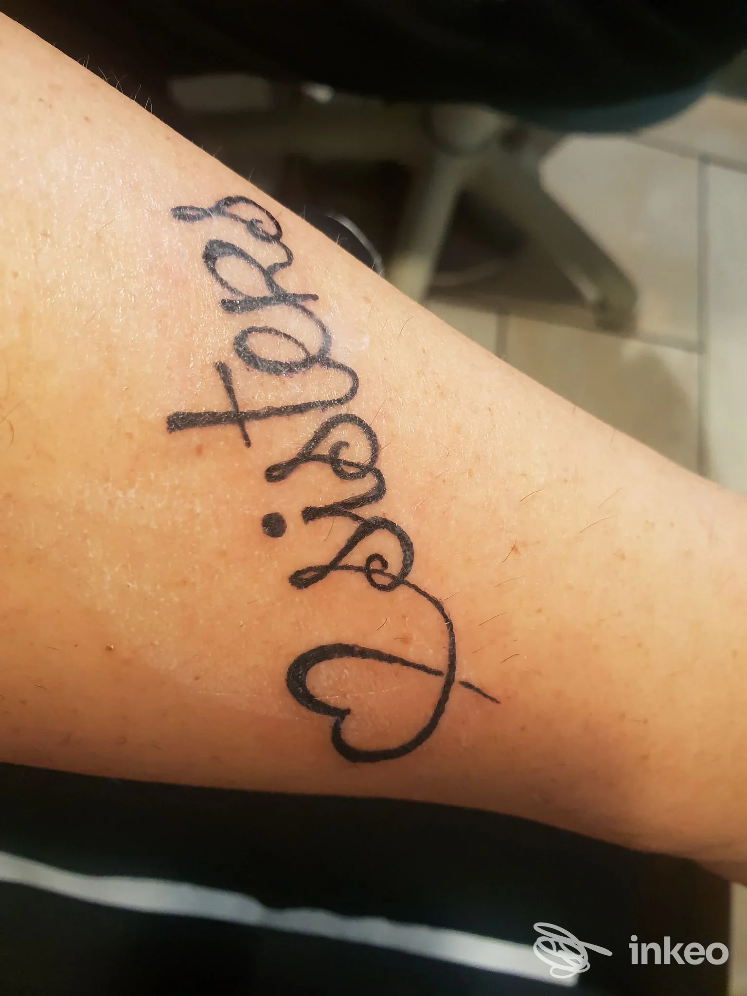 Tattoo estetico su avambraccio: scritta corsiva verticale con cuore stilizzato visibile sulla pelle, senza volti o elementi di interfaccia.