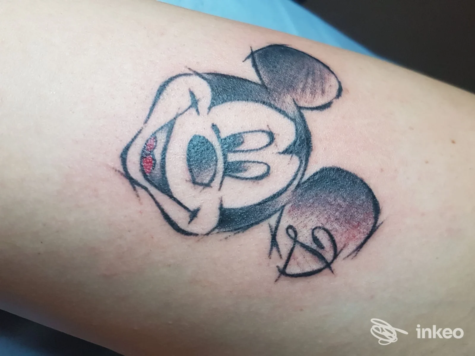 Tattoo su braccio raffigurante una testa di topo stilizzata con orecchie rotonde, contorni neri e sfumature di colore; disegno su pelle visibile.