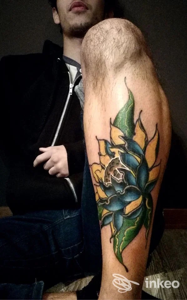 Foto di braccio destro di un uomo con tatuaggio colorato a foglie verdi, blu e gialle; il volto è parzialmente visibile sullo sfondo, senza elementi di social media o testo sovrapposto.
