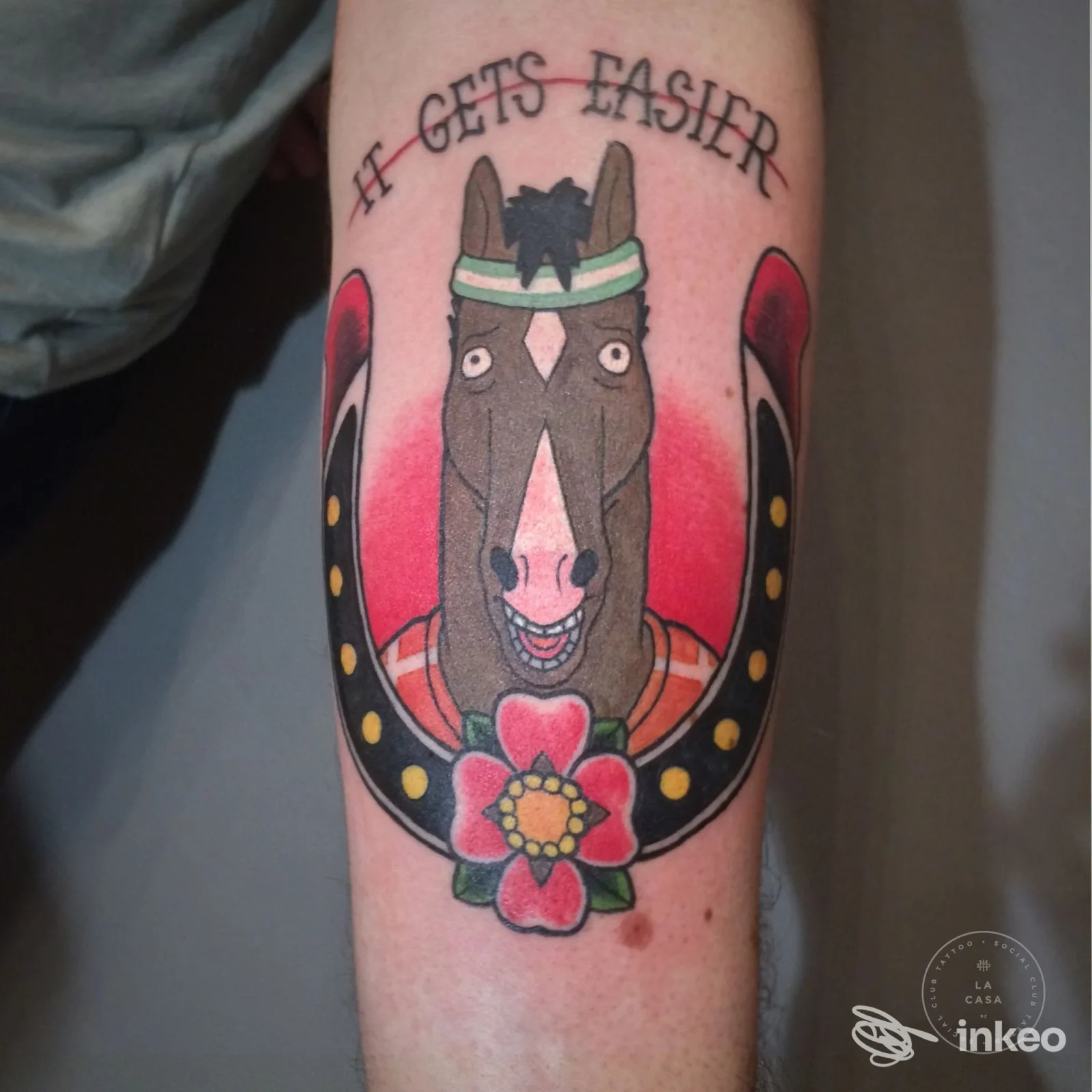 Tatuaggio colorato di un asino stilizzato con collarino verde, gancio a ferro di cavallo e fiore rosa sull'avambraccio, senza presenza di volto.
