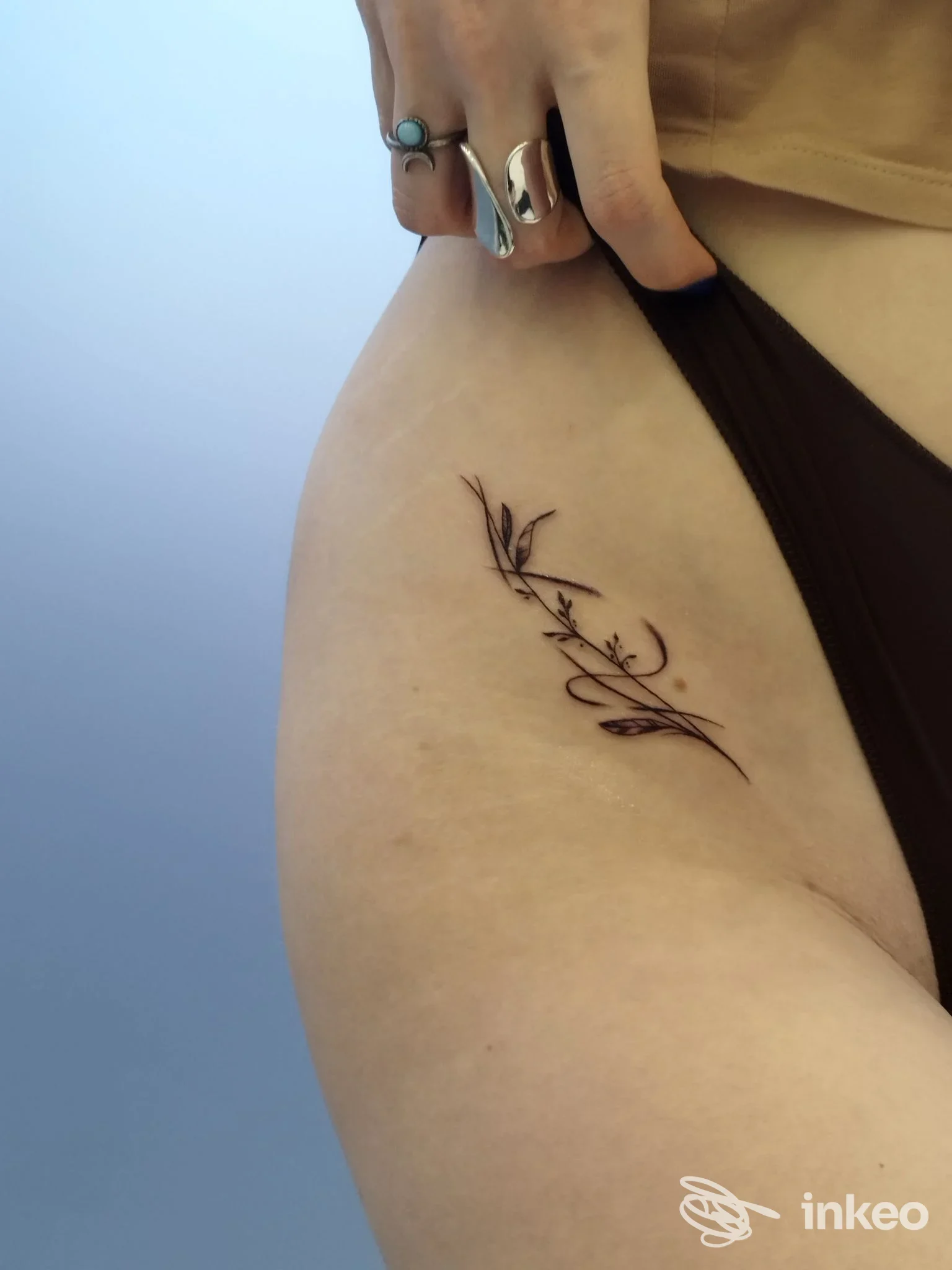 Tattoo a linea sottile su pelle, raffigurante rami o foglie, posizionato sulla coscia/anca sinistra di una persona, con bikini visibile.