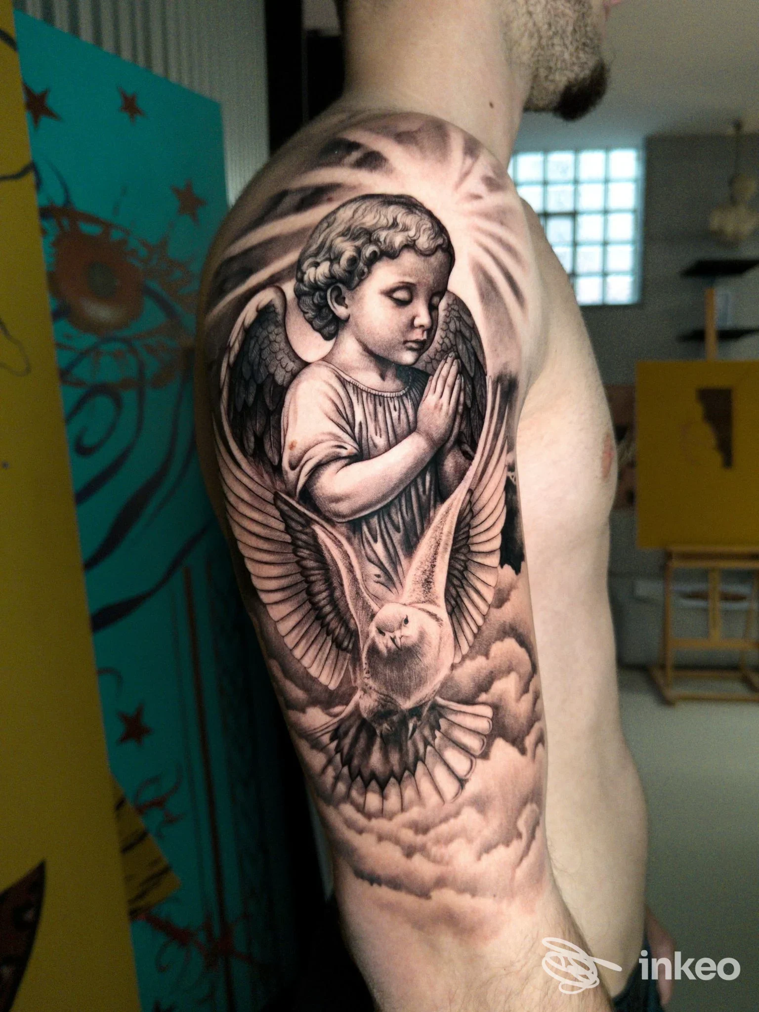 Tattoo realistico su braccio raffigurante un angelo alato che prega, con una colomba tra nuvole; pellechiamo la pelle umana come supporto principale nello studio di tatuaggi L'Arte del Tatuaggio.