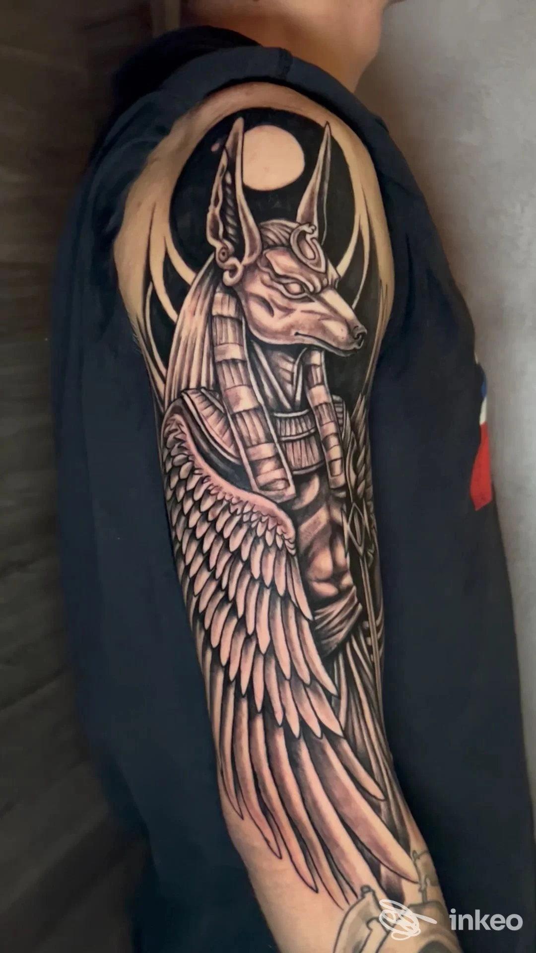 Tattoo winged creature su braccio, tonalità scure, contorni netti e sfumature nero-grigio su pelle reale. Ritratto di braccio senza viso visibile.