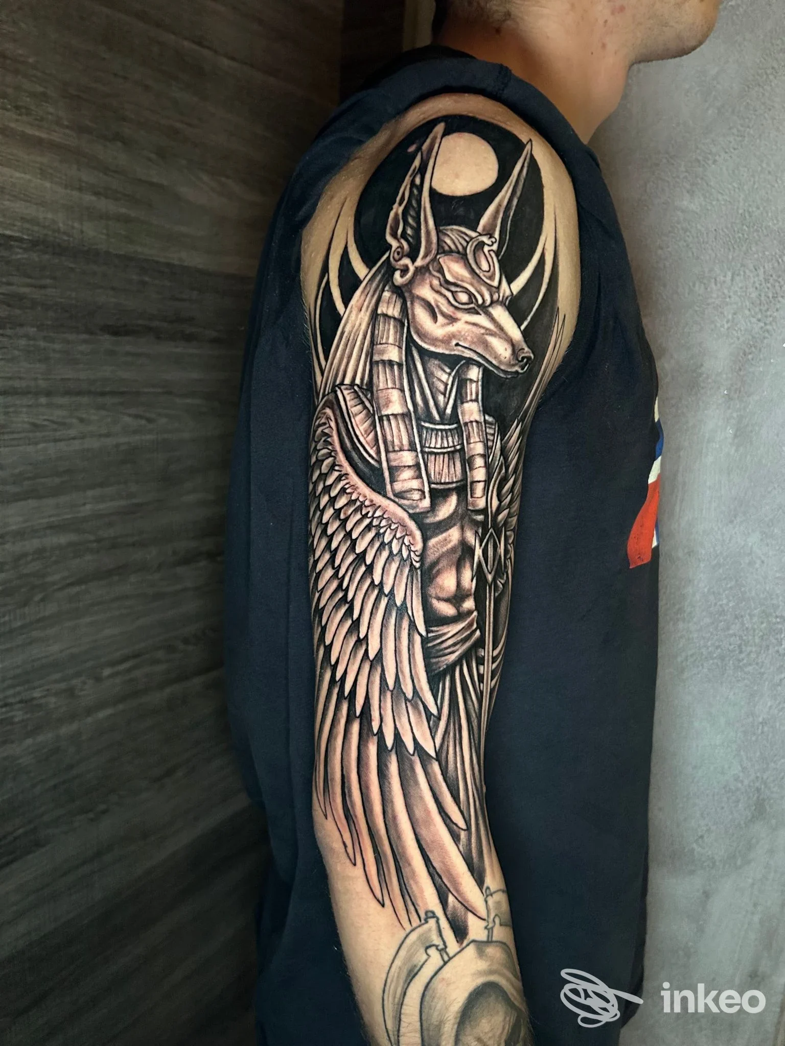 Tattoo realistico su pelle di braccio raffigurante Anubi con ali, eseguito in tonalità nere e grigie, visibile sul lato esterno del braccio.